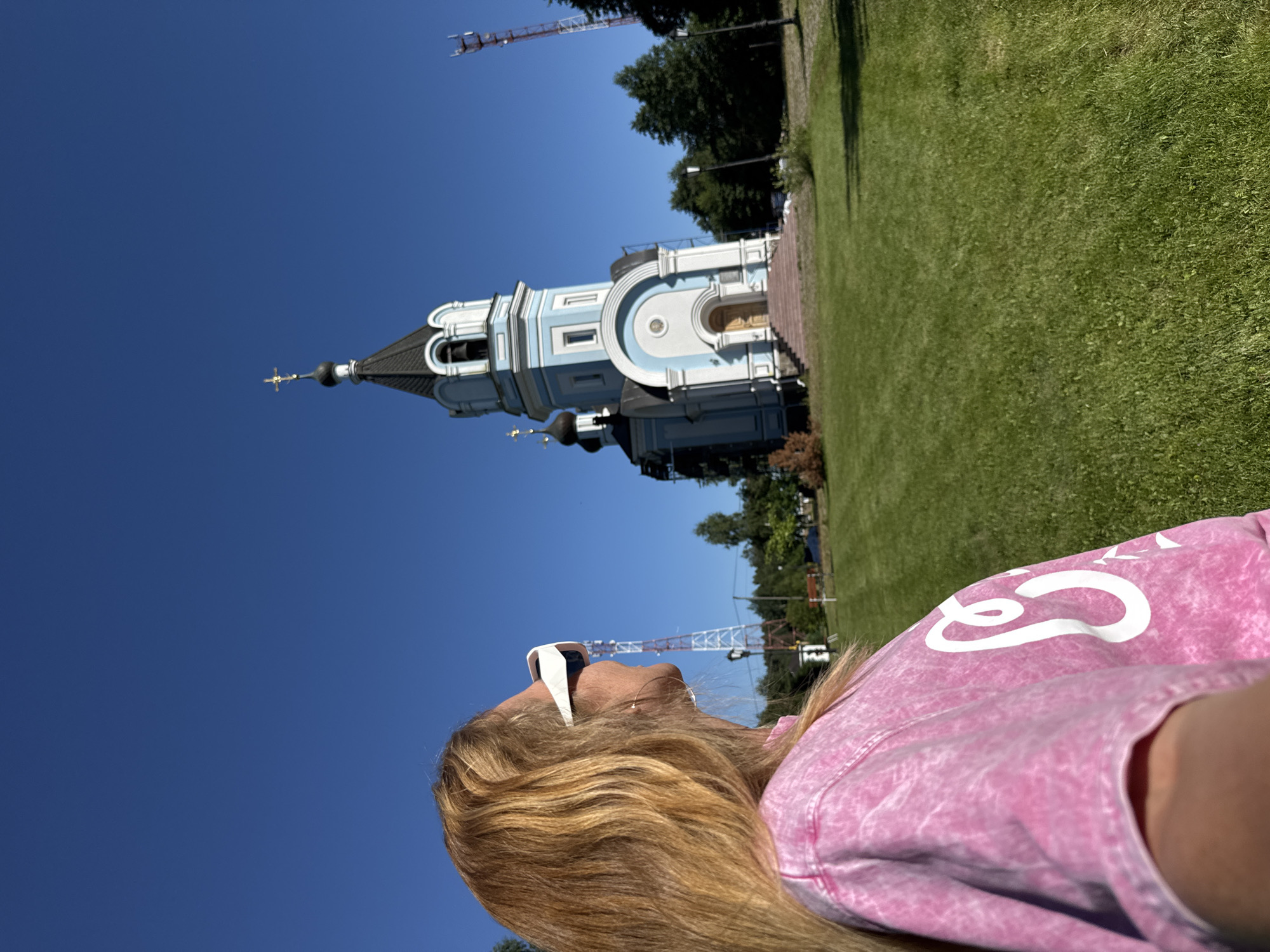 An outdoor, eye-level, medium shot shows a blonde woman from the shoulders up, facing right and looking toward a blue and white domed church in the background. The woman is wearing a pink tie-dye t-shirt and white-rimmed sunglasses. Her blonde hair is shoulder length. The church, in the middle ground, has a tall bell tower topped with a gold cross and a black, onion-shaped dome. The church’s main dome is black and also topped with a gold cross. A red and white cell tower is visible to the left of the church, and a dense line of trees forms the background. The scene is set on a bright, sunny day with a clear blue sky, and the foreground is a grassy lawn.