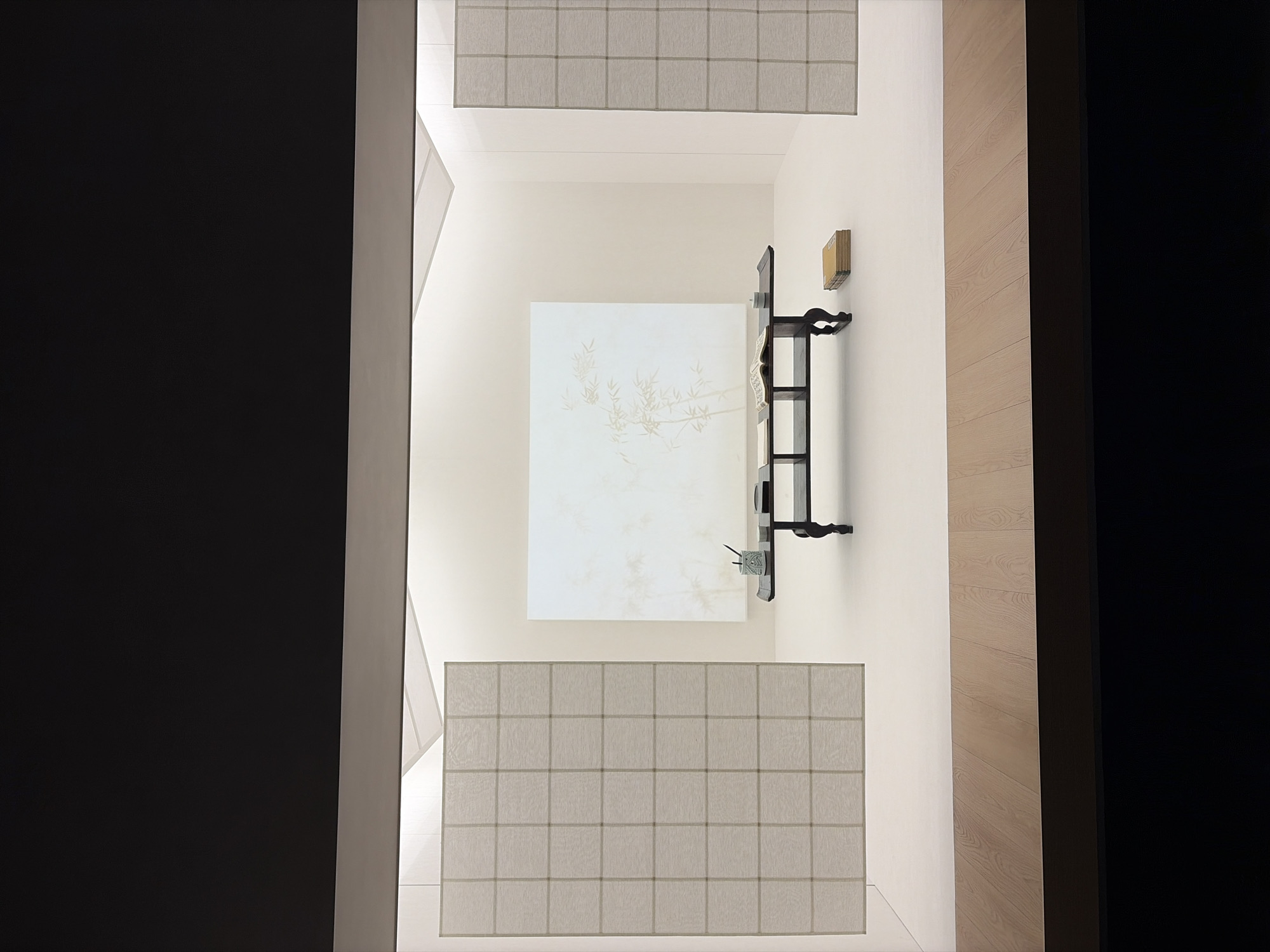 This is a photograph of an interior scene, likely an art installation or a meticulously staged display. The environment is minimalist and serene, dominated by neutral tones and clean lines. The foreground features a light-colored wooden floor. The midground reveals a white, expansive wall with a large, pale artwork depicting bamboo branches. In front of this artwork, a dark, low table holds a few objects, including an open book, a small cylindrical container with brushes or pens, and a white ceramic dish. To the right of the table, a stack of yellow-covered books rests on the floor. The sides of the room are adorned with vertical, grid-patterned screens that diffuse light, creating a soft glow. The ceiling is angled and illuminated, contributing to the overall sense of spaciousness and tranquility. The scene appears to be set within a museum or gallery space in Seoul, South Korea, possibly at the National Museum of Korea or a contemporary art institution, given the style and presentation. There are no people visible in the image.