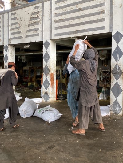 In Shurandam, Afghanistan, an industrial setting depicts men carrying heavy white sacks. The men wear traditional Afghan clothing. The building shows a two-story structure with striped and diamond pattern decor. Several white bags rest on the floor. The floor shows scattered debris.