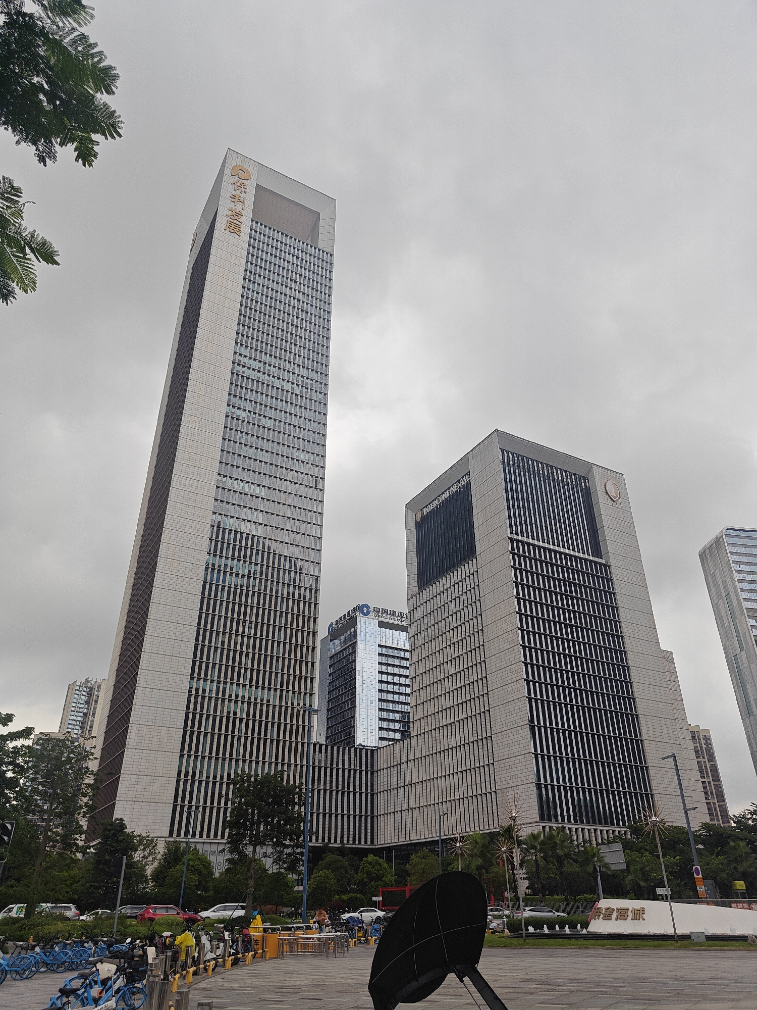 The image shows a street-level view of modern skyscrapers in the Shunde District of China. The sky is overcast. In the foreground, a paved area is visible with a black parabolic-shaped object mounted on a pole in the center. To the left, rows of blue bicycles are parked. Further back, there is a street with several cars, including red and white vehicles. Trees and green spaces are interspersed between the buildings.
The prominent buildings include a tall, slender skyscraper with golden Chinese characters on its upper section, and a wider, rectangular skyscraper to its right, featuring the "InterContinental" logo. Another glass-fronted building is visible between these two. In the distance, other high-rise buildings can be seen. A large white structure with golden Chinese characters, possibly a landmark or sign, is situated in the mid-ground on the right side of the image. People are visible near the parked bicycles and on the street, some appearing to be walking or standing. The overall impression is of a bustling urban environment.