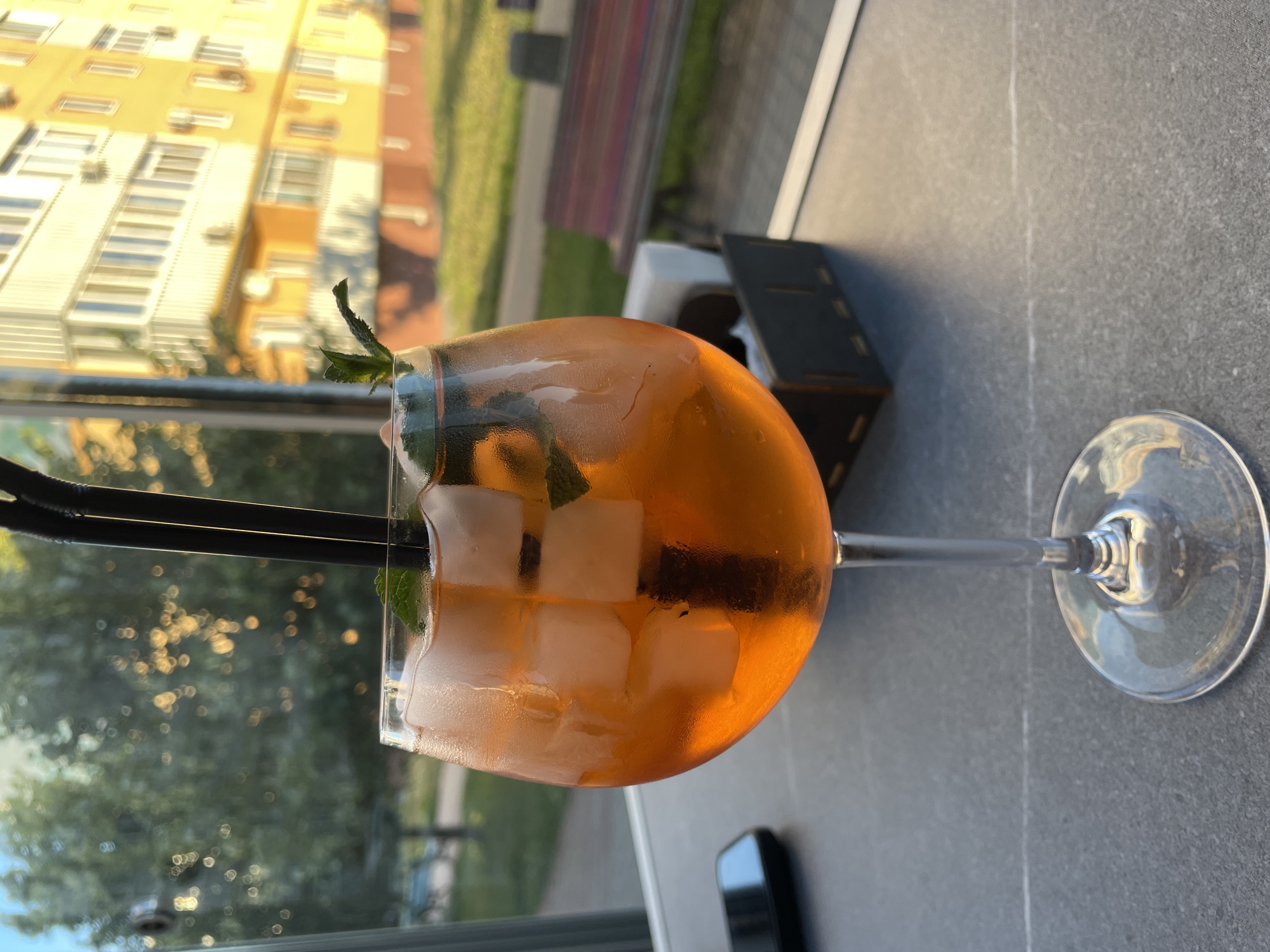 A close-up, eye-level shot shows a stemmed glass filled with an orange beverage, ice cubes, and mint leaves. Two black straws extend from the drink. The glass sits on a grey, speckled surface. In the background, through a window, a blurred green tree is visible, along with a bright yellow building with many windows and air conditioning units. A red and brown striped bench sits on a paved area outside. The scene appears to be an outdoor seating area in the city of Rozsoshentsi, Ukraine. A black mobile phone is also visible on the table to the left of the glass. A wooden holder with napkins is positioned behind the glass.