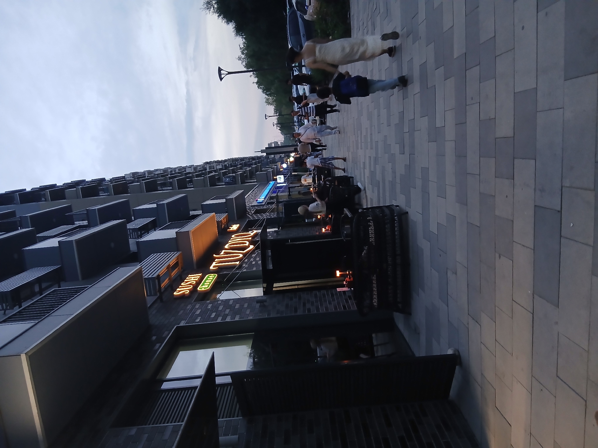 This is a vertical, outdoor shot taken at dusk, with a hazy, overcast sky. The scene depicts a pedestrian walkway in front of a modern, multi-story building. Several people are visible walking along the sidewalk. The building has a dark brick facade with many rectangular windows and balconies. A neon sign on the building reads "SUSHI" in orange and green letters, and another sign above it displays "TIZO" in orange neon. The sidewalk is paved with large, rectangular grey tiles laid in a staggered pattern. A few streetlights are present, illuminating the walkway. The overall mood is casual and relaxed, suggesting a public space where people are out for an evening stroll or heading to a nearby establishment.