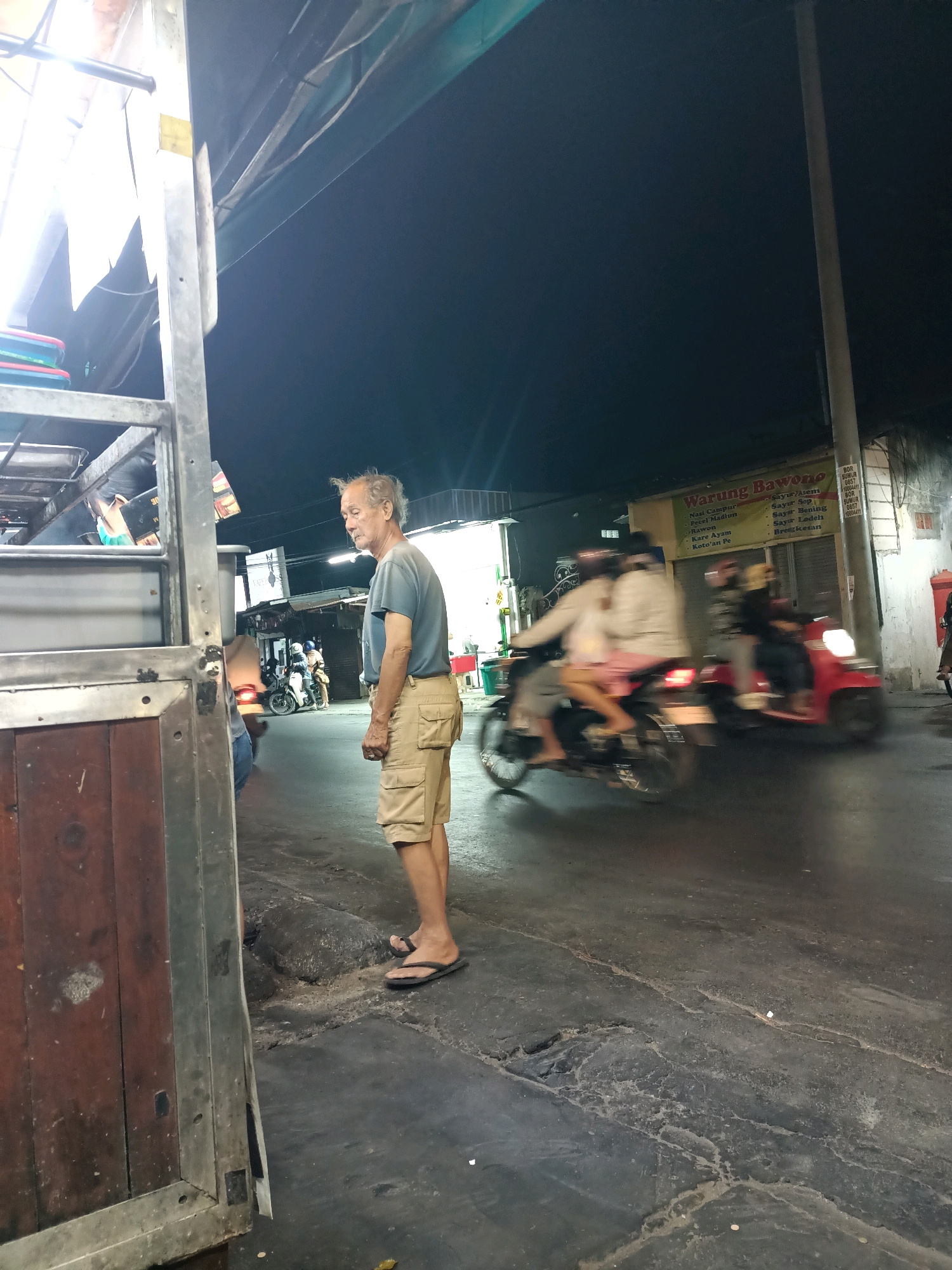 An elderly Asian man stands on a paved street at night in Surabaya, Indonesia. He is wearing a blue t-shirt and beige cargo shorts, with black flip-flops. His hair is unkempt and styled to the side. He is looking to his left. To his right, two motorcycles are moving, one red and one gray. The red motorcycle is driven by a person wearing a yellow hijab and a black top, and a passenger wearing a light-colored top. The gray motorcycle is ridden by a person wearing a white helmet and a light-colored top. Behind them, a building with a yellow sign that reads "Warung Bawono" is visible. The street is cracked and uneven. In the foreground, to the left, is the side of a metal and wooden structure, possibly a food cart or stall. The sky is dark, indicating nighttime.