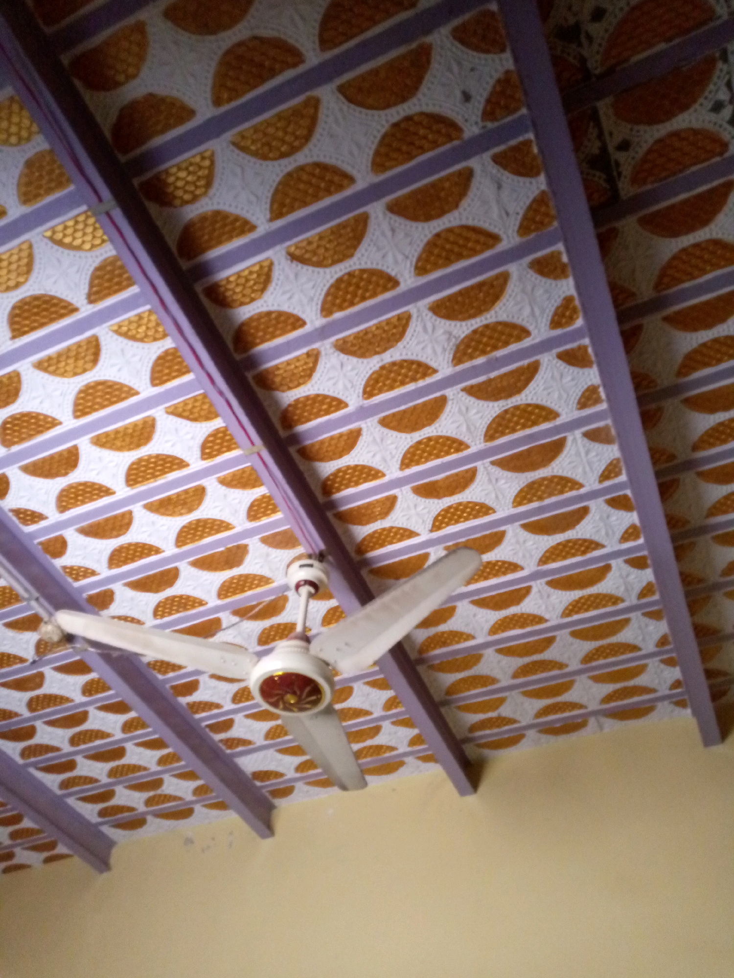 The image shows a ceiling fan mounted on a decorated ceiling. The ceiling has a pattern of alternating horizontal purple and white stripes. The white stripes contain a repeating pattern of golden semi-circles with a textured fill, resembling a basketweave or cross-hatch design. The ceiling is supported by purple beams. The ceiling fan is white with three blades and a central motor housing that appears to have red and gold accents. The lower portion of the image shows a plain, light yellow wall. This scene is located in the city of Khuzdar, Pakistan.