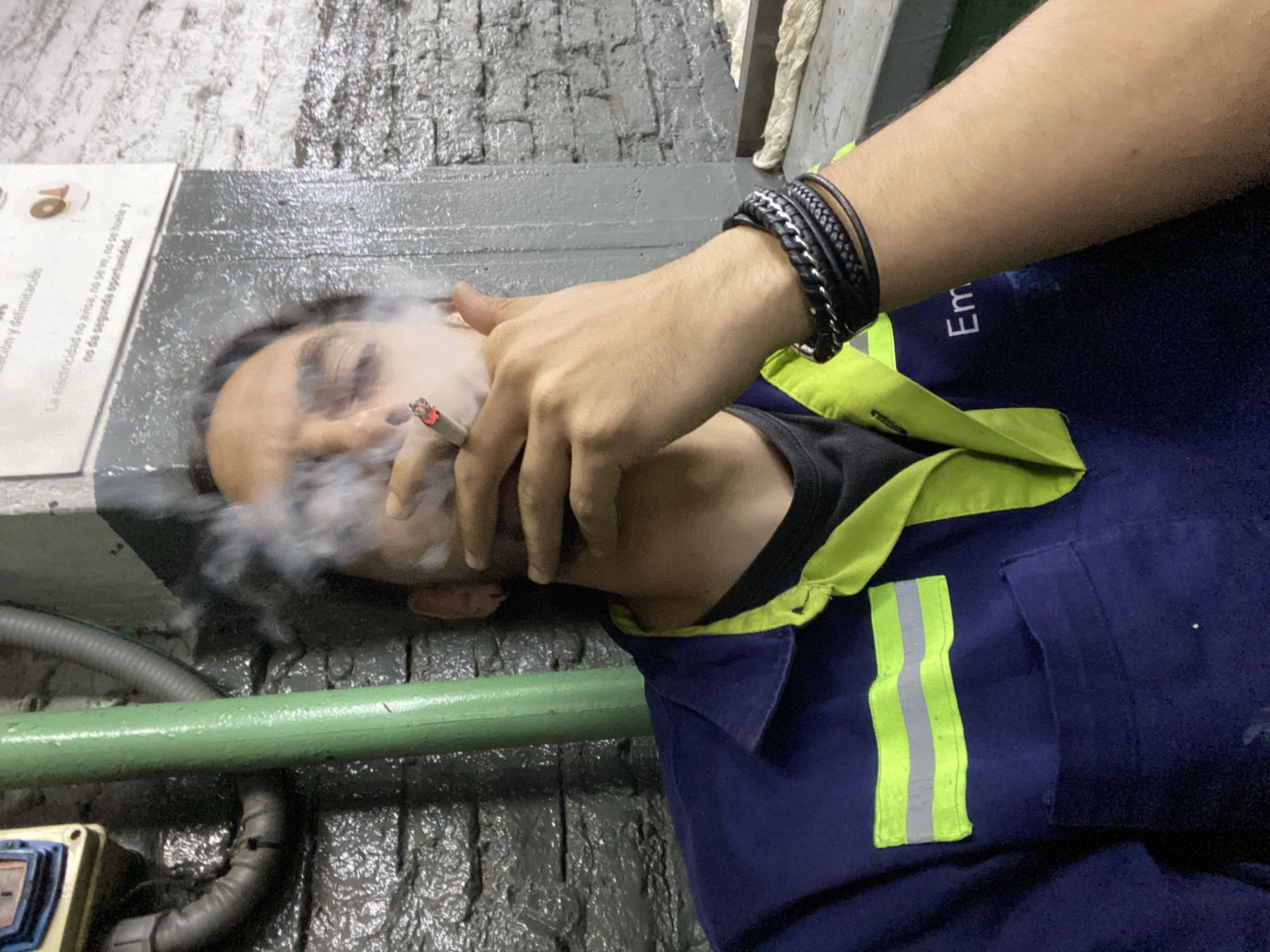 A man is seen lying down, smoking what appears to be a cigarette. He is wearing a dark blue work shirt with a neon yellow/lime green collar and stripes on the shoulder. He is also wearing a black wristband. The background appears to be an industrial setting with gray concrete walls and pipes. A sign is visible on the wall, with a text in Spanish about electricity, which suggests this location is in a Spanish-speaking area, possibly in Buenos Aires, Argentina.