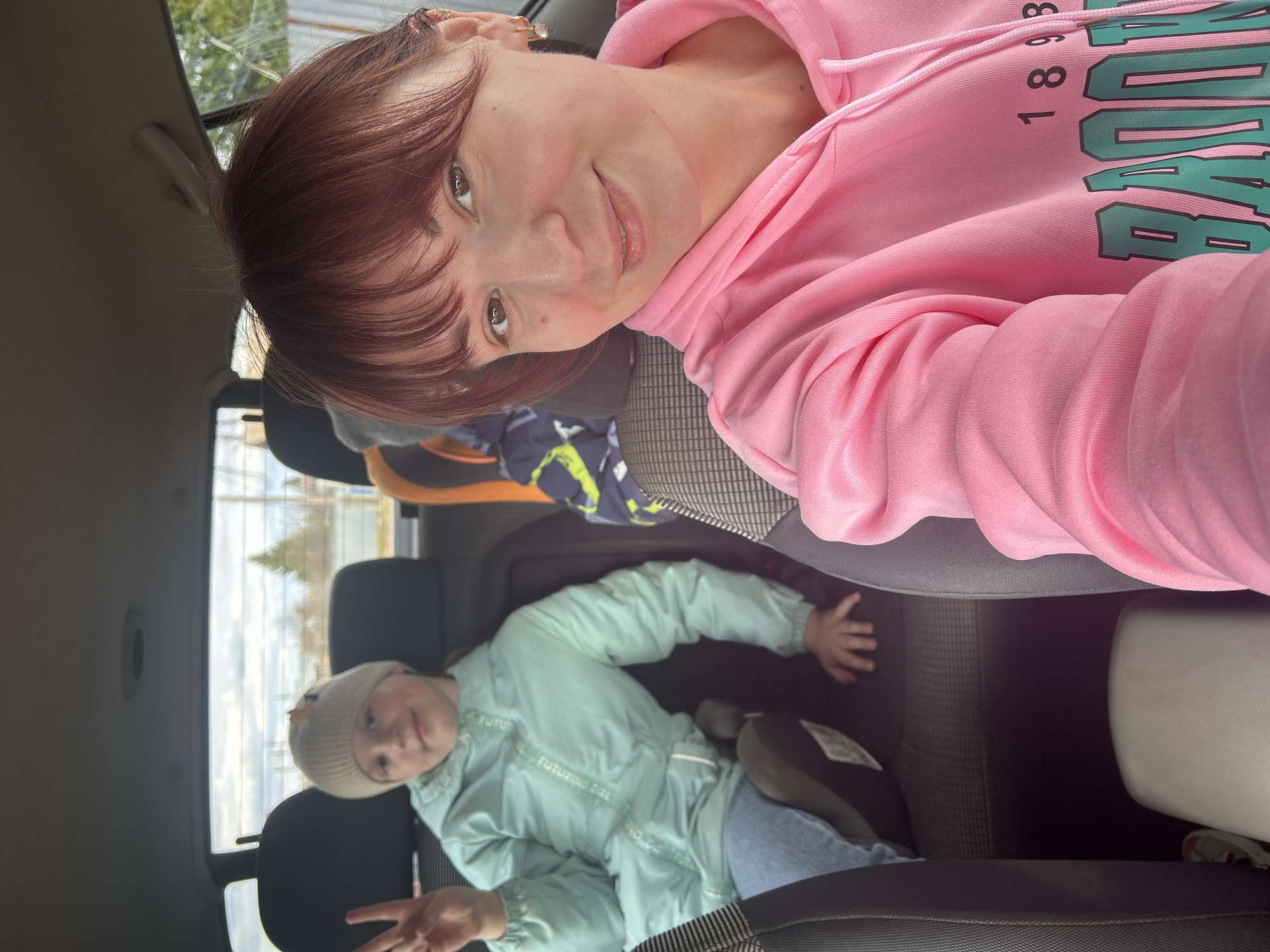 A woman in a pink hoodie and a girl in a light green jacket are in a car. The woman is in the front seat, turned to face the camera, and smiling. The girl is in the back seat, making a peace sign. Through the car windows, there is a glimpse of an outdoor area, showing a building and some trees, with an overcast sky. The location is Фоки, Russia.