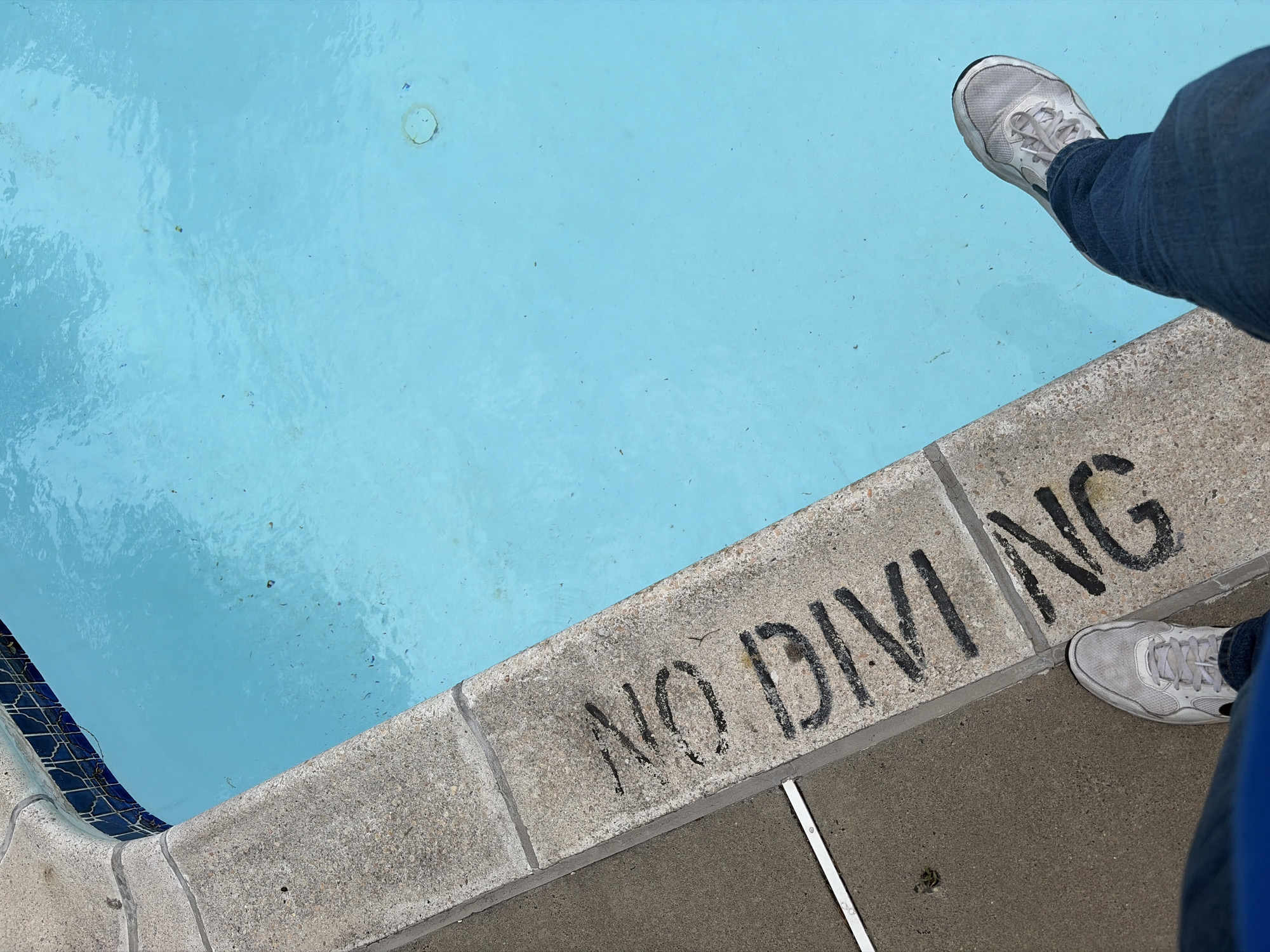 The image shows the edge of a swimming pool with a "NO DIVING" warning painted in black on the concrete border. A person wearing blue jeans and gray sneakers is standing at the pool's edge. The pool water is blue. The location is the city of Edison, United States of America.