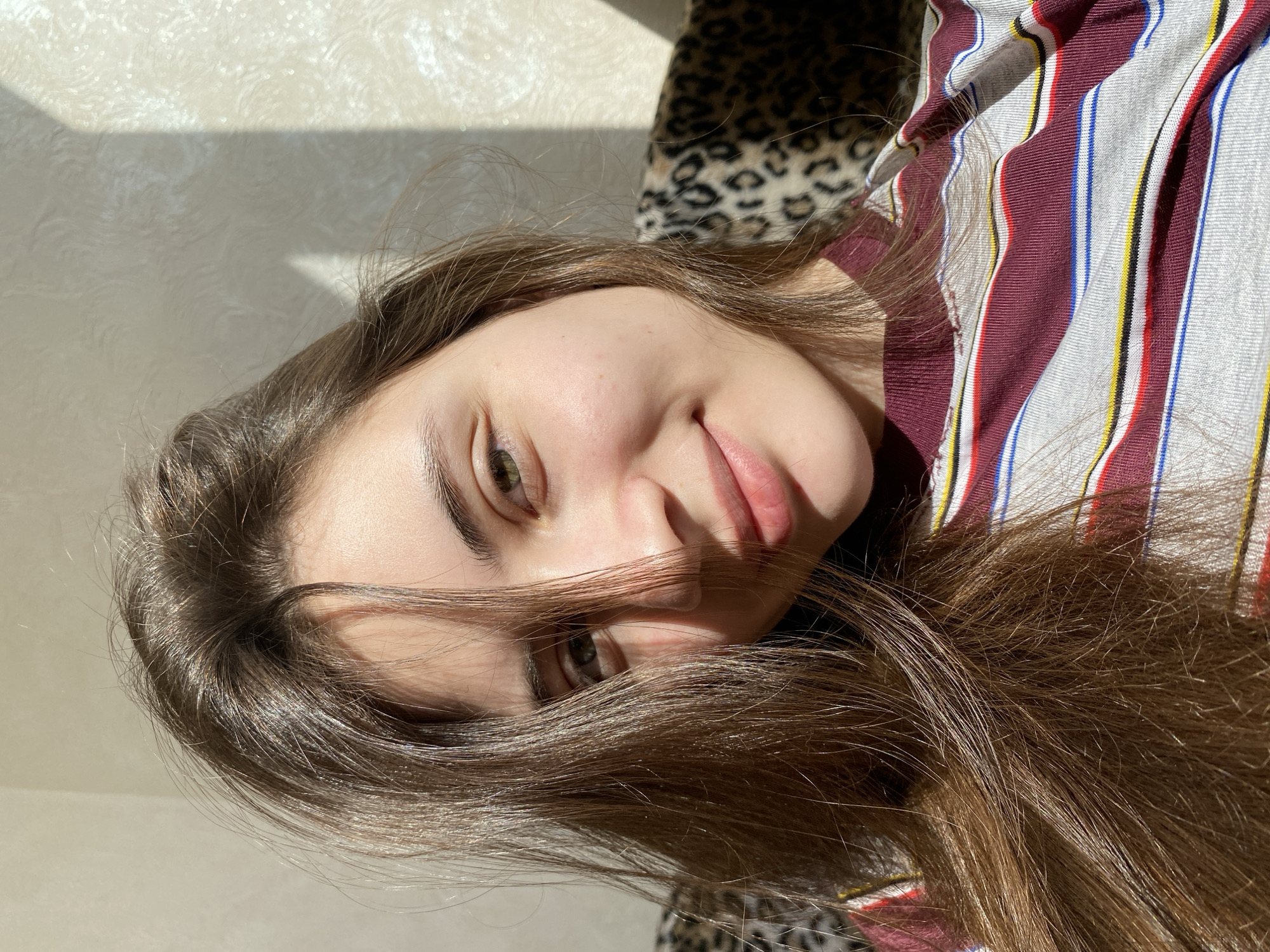 A close-up, high-angle portrait captures a young person, likely female, with long, glossy brown hair, in an indoor setting in Votkinsk, Russia. The individual is positioned so their head is tilted, looking directly at the camera with a subtle, relaxed expression. Their eyes are a striking greenish-brown, and a faint, natural smile plays on their lips. Strands of her long hair fall across her face and forehead.
She is wearing a striped shirt featuring vertical stripes in shades of maroon/dark red, white, blue, and yellow. Bright natural sunlight streams in from an unseen window, strongly illuminating her hair and face, creating highlights and casting distinct shadows on the light-colored, textured wall behind her. The strong light suggests it is daytime, possibly morning or late afternoon.
To the upper right of the frame, behind her head, a section of an object or surface covered in a black and brown leopard print pattern is visible, adding a contrasting texture to the scene. The lighting emphasizes the texture of her hair and the subtle patterns on the wall. There is no visible text in the image. The overall impression is casual and natural, captured in warm daylight.