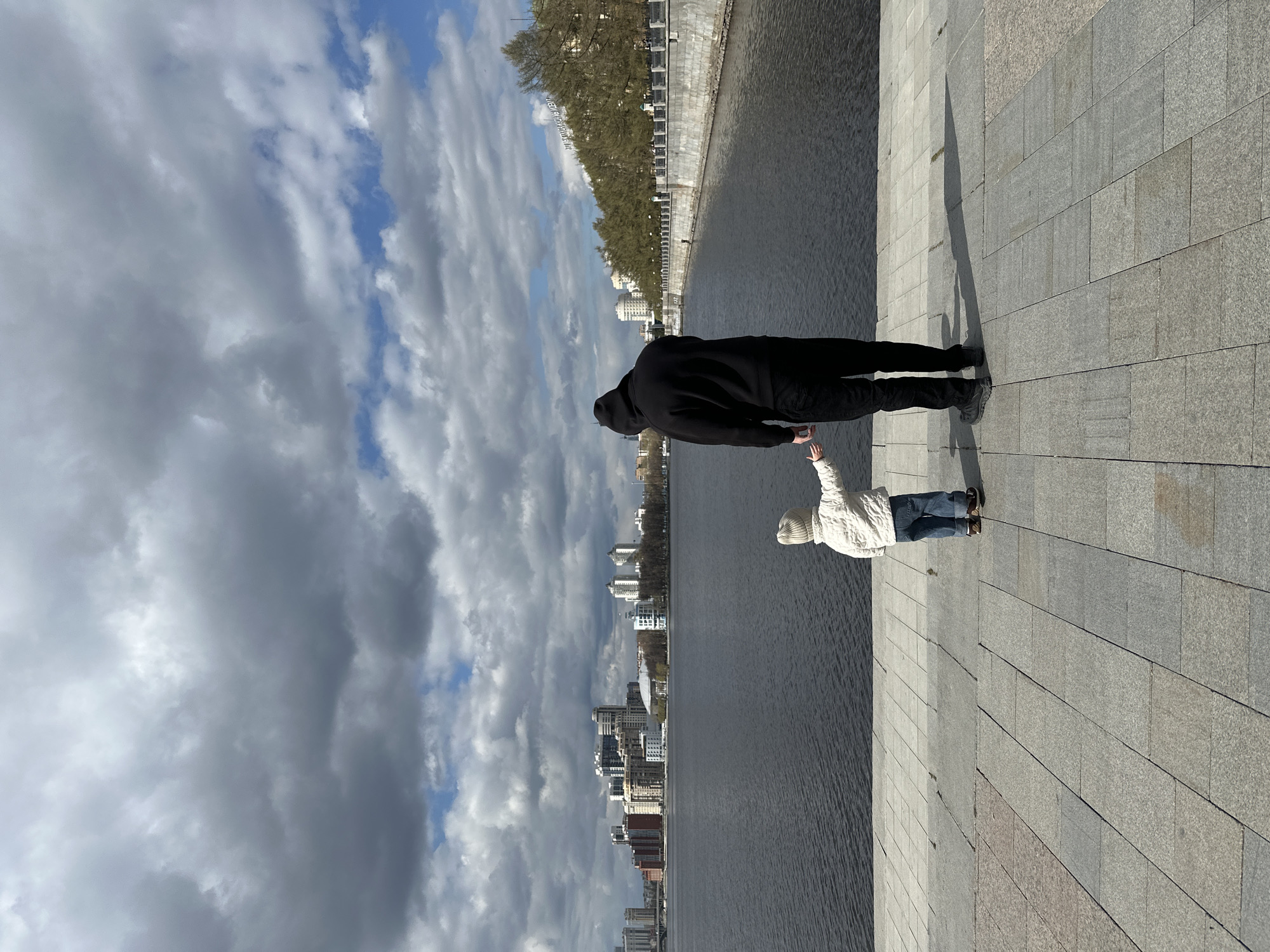 A street-level shot depicts a person in a black hoodie and pants holding hands with a child wearing a white jacket and jeans. They stand on a gray stone walkway next to a body of water. Across the water, buildings rise into a cloudy sky with patches of blue. The scene is identified as Yekaterinburg, Russia, based on architectural cues.