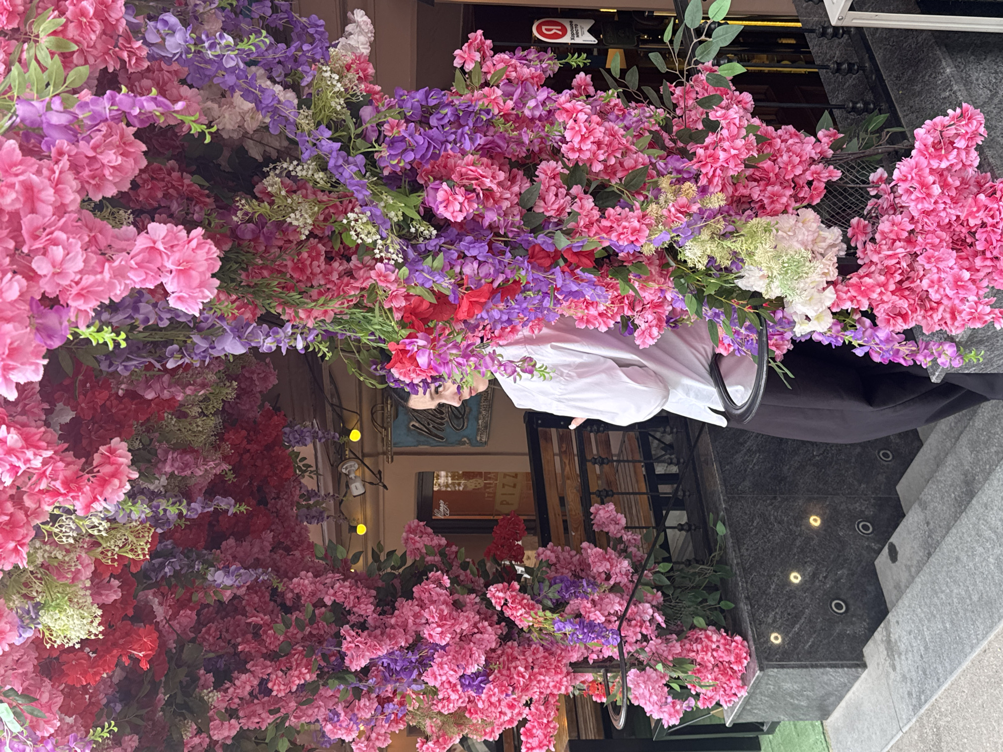The image shows the exterior of a restaurant, possibly a pizzeria, in St. Petersburg, Russia. The entrance is heavily decorated with pink, red, and purple artificial flowers. A person in a white shirt and dark pants is partially visible behind the flowers. The facade of the building is decorated with a sign that says "PIZZA" and "Village ITA". The entrance has several steps up to the restaurant. The sidewalk is visible at the bottom right of the image.