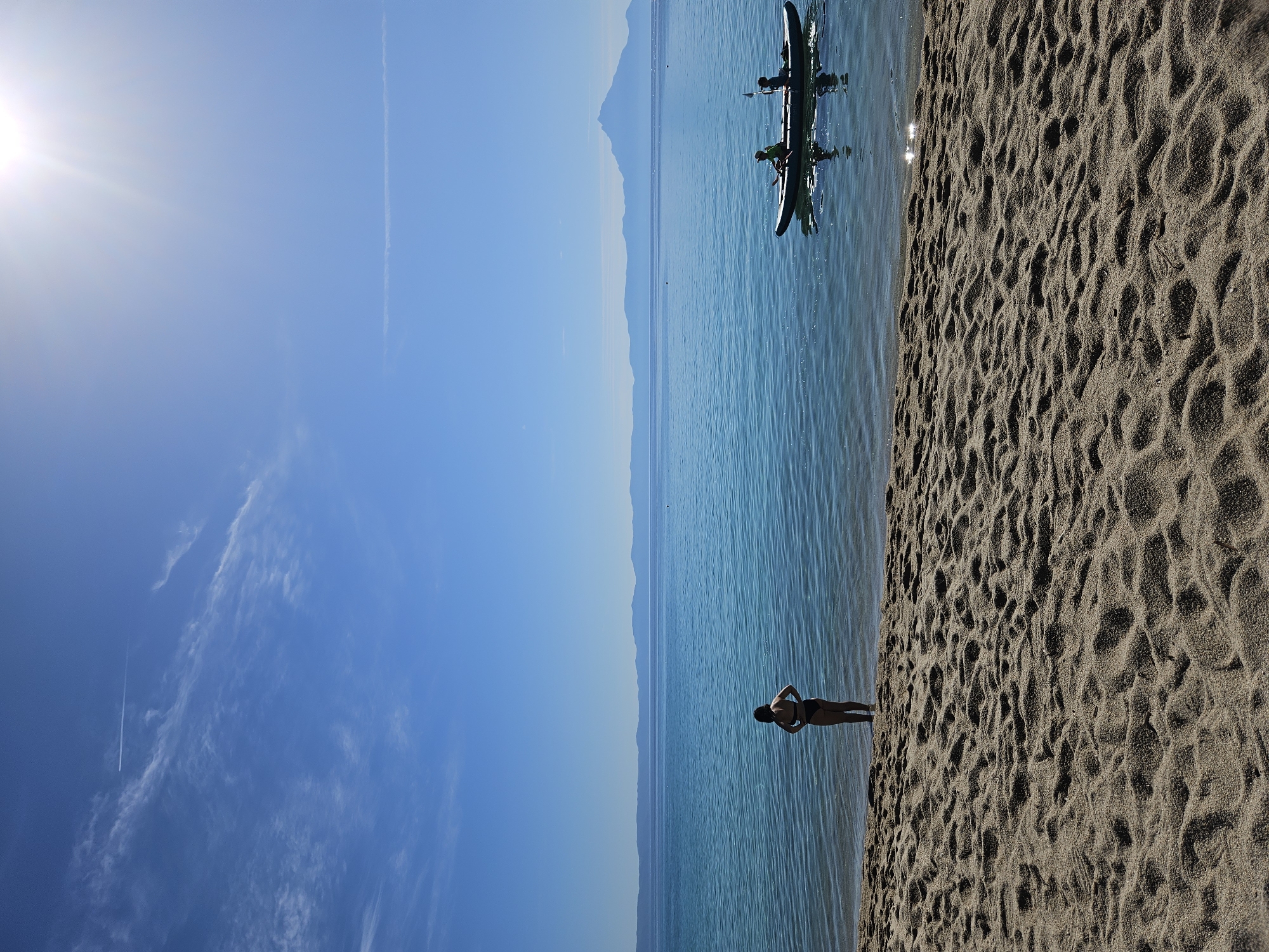 The image depicts a tranquil beach scene on a sunny day in Toroni Municipal Unit, Greece. In the foreground, textured sand covers the beach, leading to the edge of the clear, turquoise water. A person wearing a dark bikini stands at the water's edge, facing the sea with their back to the camera. Further out on the water, a small kayak carries two individuals, one holding a paddle. The background is dominated by a vast expanse of calm sea, with a distant, hazy coastline featuring mountainous terrain. The sky is a brilliant blue, punctuated by scattered white clouds and the bright sun with visible rays in the upper right corner. An airplane trail is visible as a thin white line across the sky, indicating flight activity. The overall impression is one of peaceful natural beauty and recreational activity by the sea.