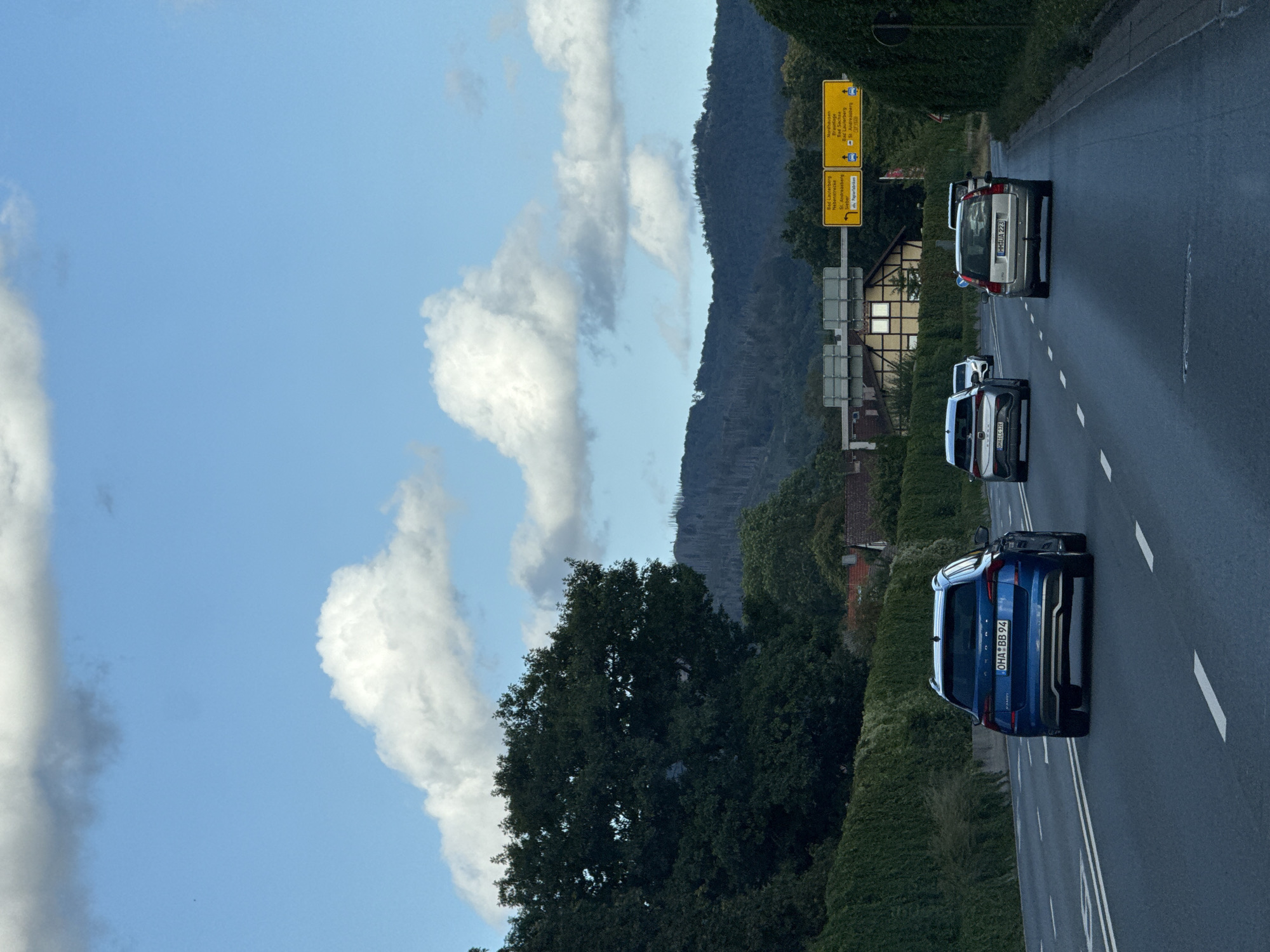 This is a daytime, eye-level, vertical shot taken from a moving vehicle on a highway. The road has multiple lanes marked with white lines. In the foreground, several cars are driving on the highway. The closest car is a blue SUV, followed by a white SUV, a silver SUV, and another white car further back. The highway is lined on both sides by dense green hedges. In the background, there are buildings with yellow signs indicating directions to various towns, including Herzberg, Bad Lauterberg, and Nordhausen. Beyond the buildings, there is a dark, wooded hillside under a bright blue sky with scattered white clouds. The overall impression is of a scenic drive through the German countryside.