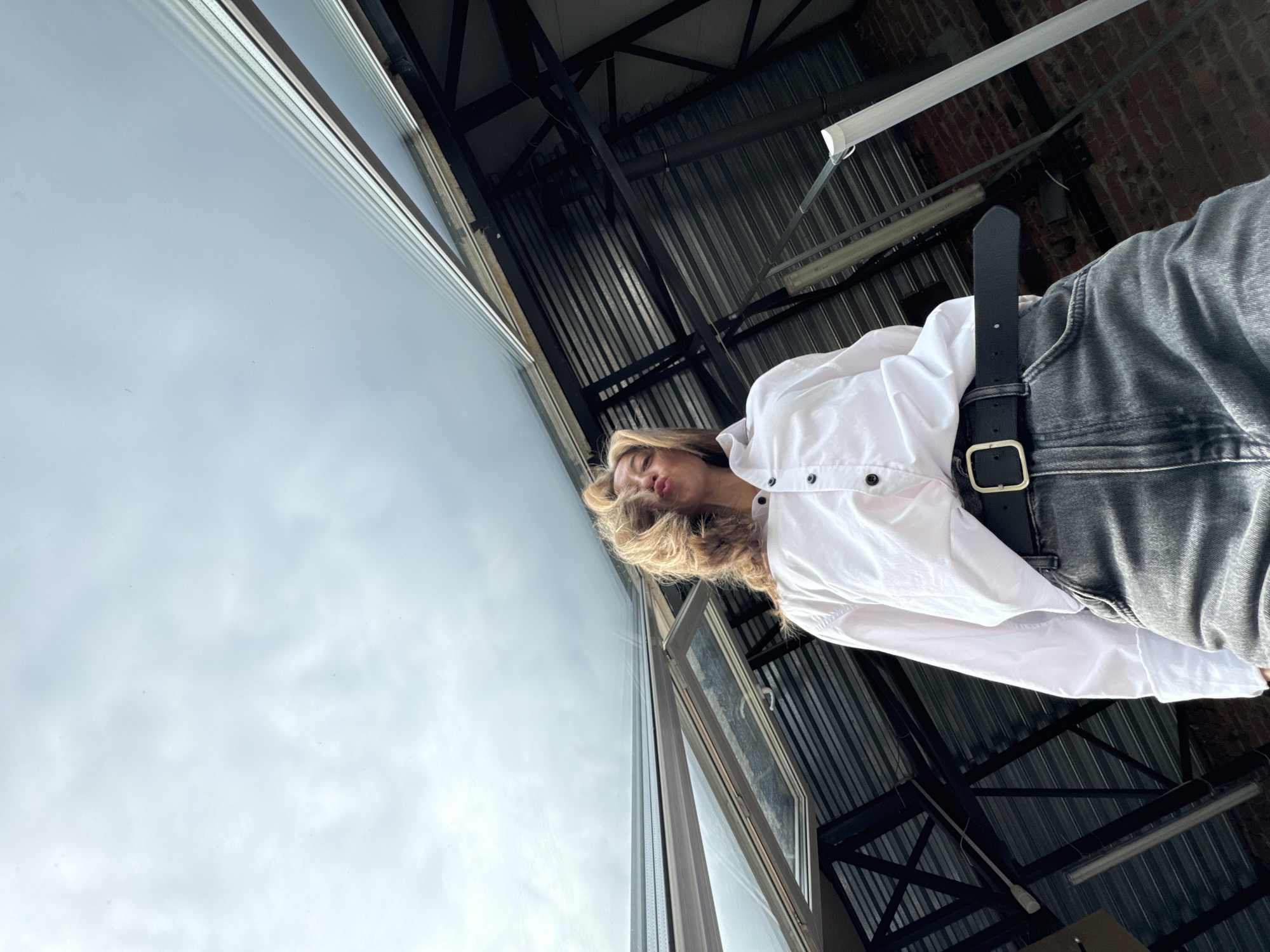A low-angle, medium shot captures a person from the waist up, looking up at the camera with their lips pursed. They are wearing a white button-down shirt with dark buttons and dark wash jeans cinched with a black belt with a gold buckle. Their blonde hair is tousled and appears to be blown by the wind. The background consists of a corrugated metal roof with exposed beams and a partially opened skylight. The sky above is overcast and cloudy. The location is not discernible from the image itself, and there is no indication it is in Chaikovsky, Russia.