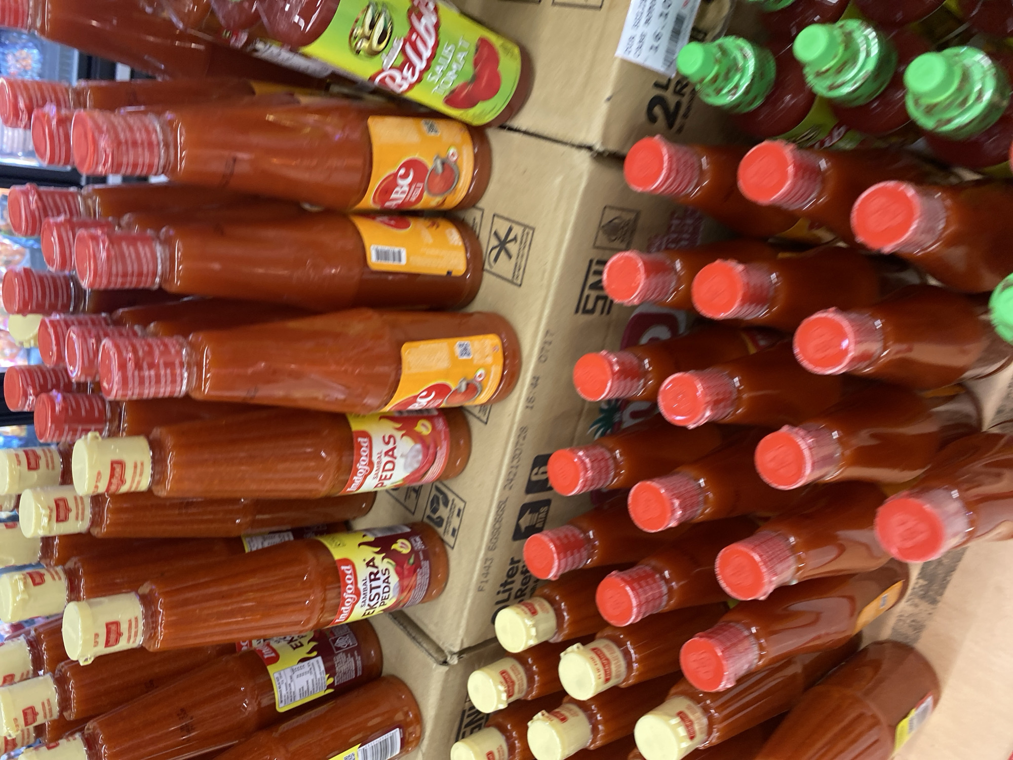 The image displays a retail setting in Banda Aceh, Indonesia, likely a supermarket or a local store, showcasing a large assortment of bottled chili sauces and ketchup. The primary focus is on numerous bottles of Indofood Sambal Ekstra Pedas and Indofood Sambal Pedas, characterized by their distinctive red sauce and yellow or white labeling. Several bottles of ABC brand ketchup are also visible, identifiable by their orange labels. Additionally, a bottle of Belibis Saus Tomat (ketchup) is present, featuring a green label. The products are arranged in rows, some stacked on cardboard boxes that bear Indonesian text and barcodes. The overall impression is one of a well-stocked product display.