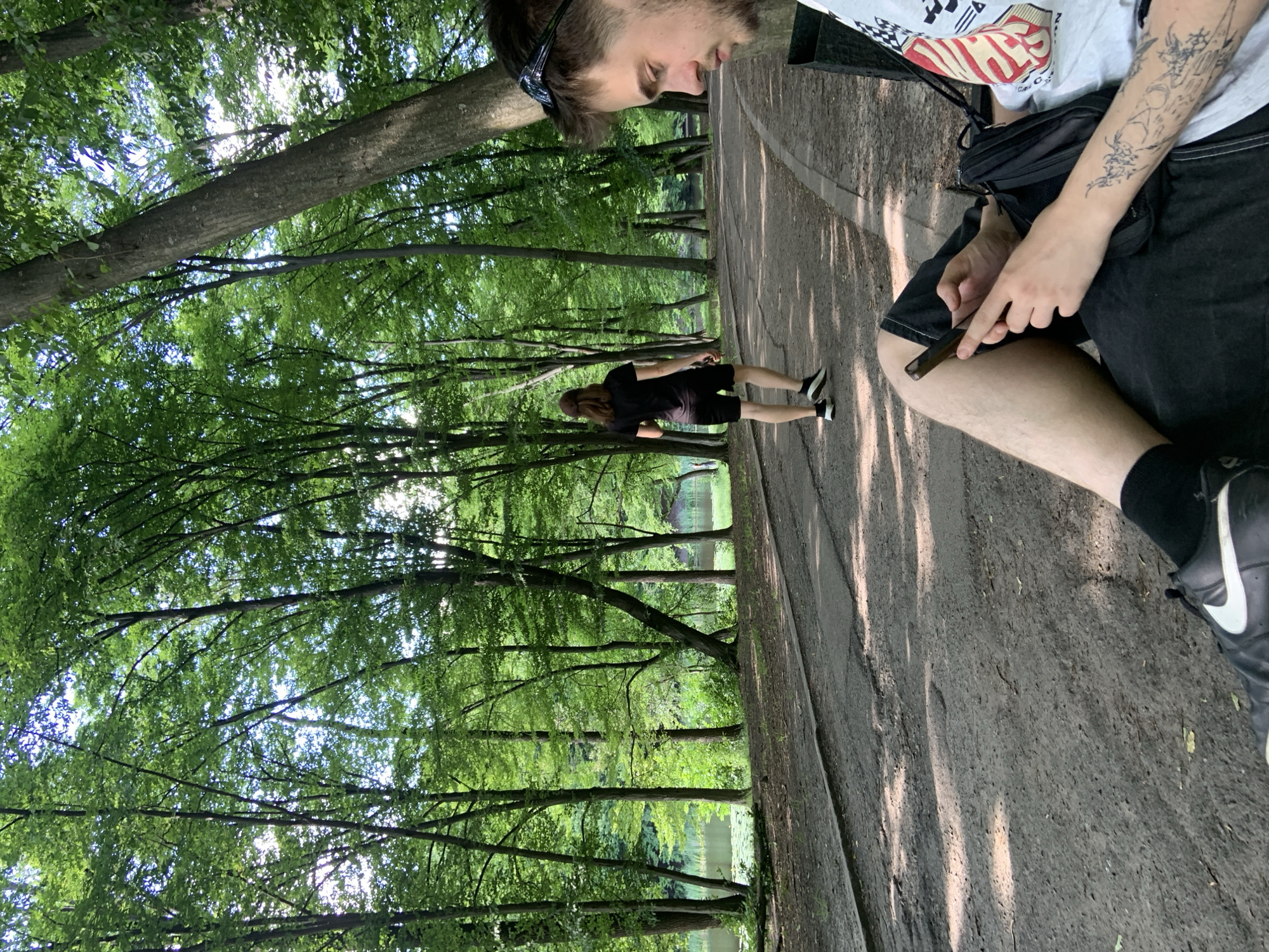 A man with dark hair and a beard is sitting on a bench in a park, looking down at his phone. He is wearing a white t-shirt with a graphic print, black shorts, and black socks with white sneakers. His left arm is visible and has tattoos. A black bag is resting next to him on the bench. In the background, a person wearing a black t-shirt and black shorts walks away from the camera on a paved path. The path is lined with trees, and a body of water with lily pads is visible to the left. The scene appears to be in a park in Kyiv, Ukraine, with lush green trees and dappled sunlight.