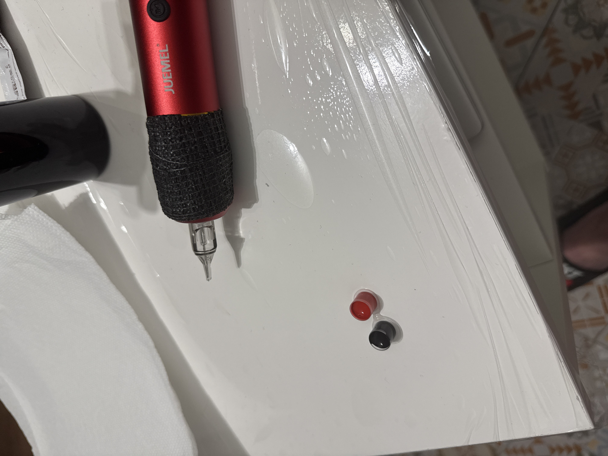 The image captures a close-up of tattooing or permanent makeup equipment laid out on a sterile white surface, likely in Sofia, Bulgaria. The main subject is a red, cylindrical tattoo machine, prominently displaying the brand name "JUEMEL" in white text. Its grip is wrapped in black, textured cohesive tape, and a clear cartridge needle is attached to its tip, ready for use.
To the right of the machine, two small, connected ink caps are visible: one red and one black, both empty and resting on the surface. The white surface itself, possibly a table or cart, is covered with clear plastic wrap, a common practice for maintaining hygiene in such settings. Several small liquid droplets are scattered across the plastic film, suggesting it might have been disinfected or sprayed.
A portion of a black, cylindrical object, possibly a power supply or another accessory, is visible on the far left. Below it, a white, disposable towel or tissue contributes to the clean setup. In the blurred background, a tiled floor with a light-colored pattern can be seen, indicating an indoor, professional environment such as a tattoo studio or permanent makeup clinic. The overall scene depicts a moment of preparation before a cosmetic or artistic procedure. No individuals are present in the image, and the time of day is not discernible.