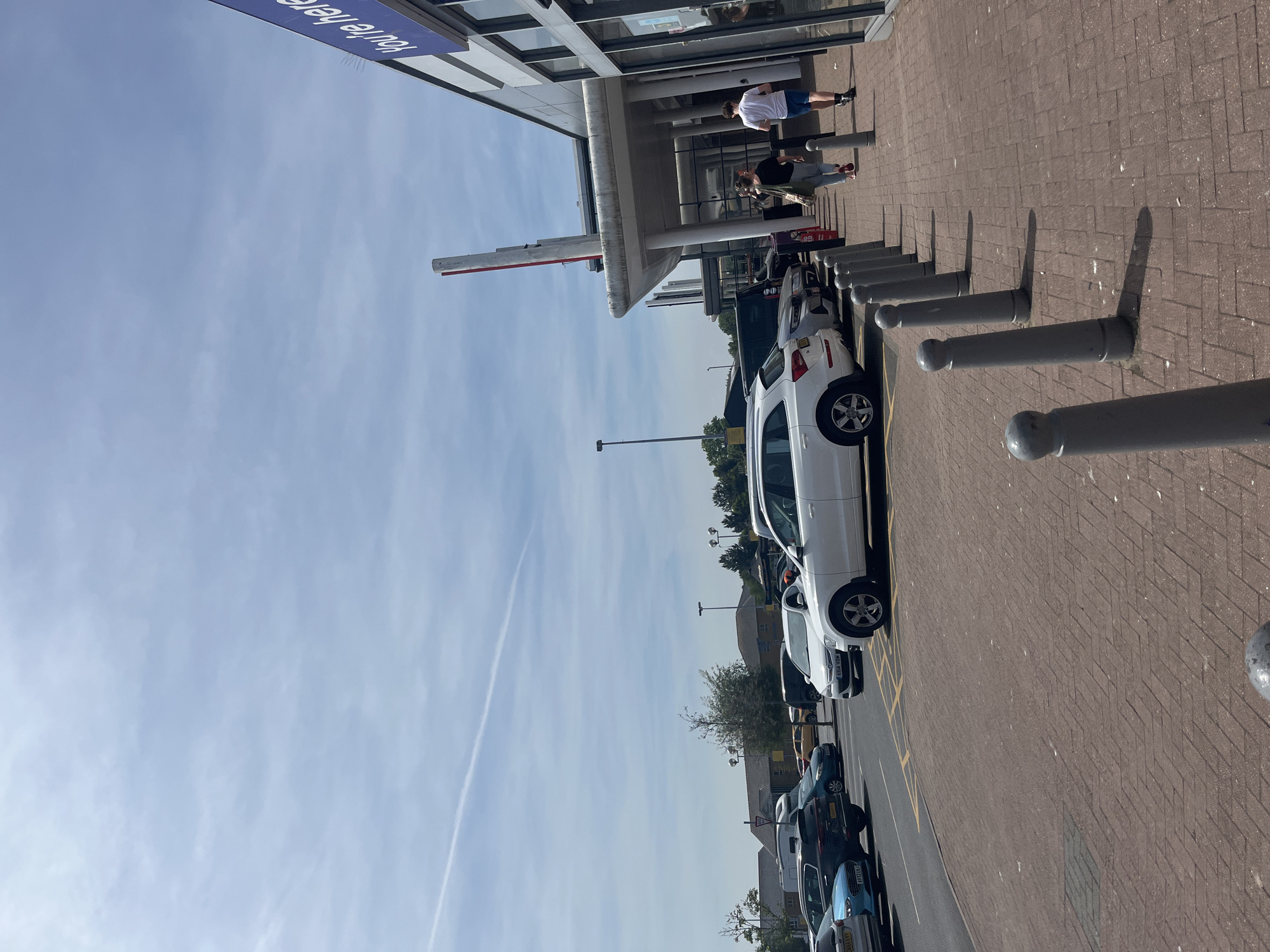 The image depicts a street scene in East Suffolk, United Kingdom. A line of parked cars is visible along a building, with a white car being the most prominent. Pedestrians walk along the paved area adjacent to the building. Bollards line the pavement. The sky is blue with some clouds. A sign reading "You're Here" is partially visible on the building.