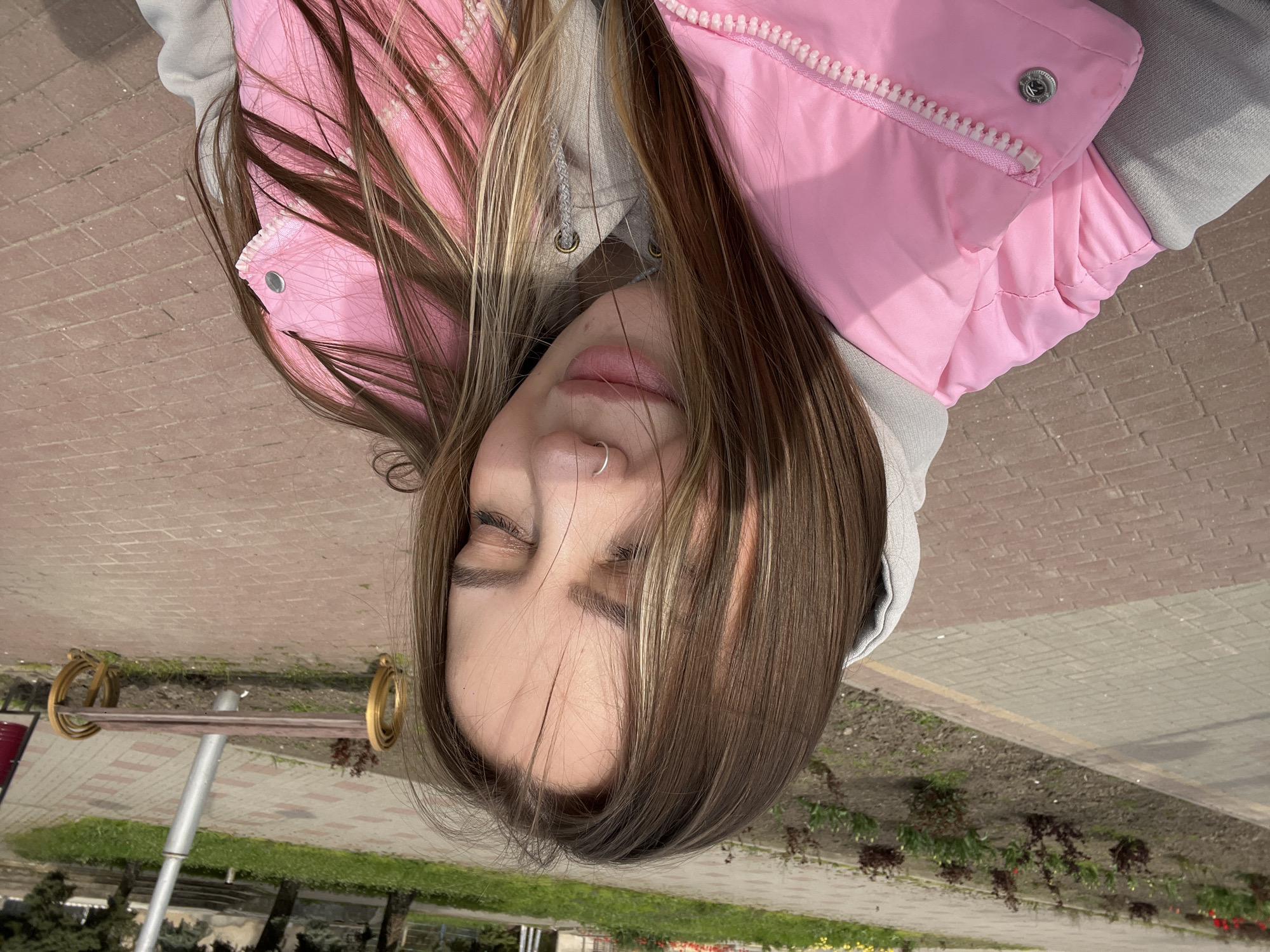 A young woman lies supine on a brick walkway outdoors in Novopokrovskaya, Russia. She wears a pink puffer jacket over a gray hooded sweatshirt. The walkway runs alongside a narrow strip of grass and plants with a park bench nearby. Her eyes are closed.