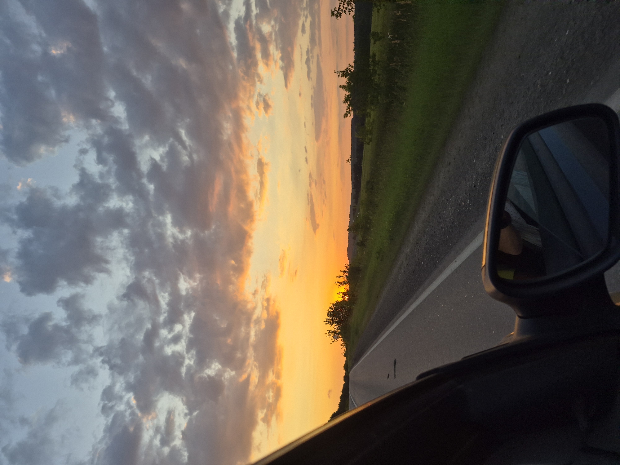 The image shows a view from the passenger side of a car traveling on a paved road during sunset. The sky is filled with a dramatic display of orange and yellow hues, with scattered dark clouds. The road curves to the left, with a grassy shoulder and trees lining the right side. In the distance, rolling hills are visible against the bright sky. The side-view mirror of the car is visible in the foreground, reflecting a portion of the interior and the passenger. The overall impression is one of a peaceful, scenic drive through the countryside of Забегалово, Russia.