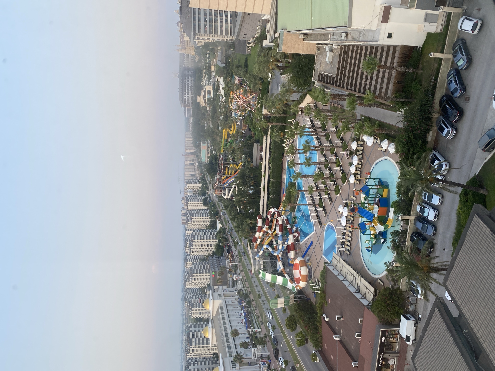 This is an aerial, slightly elevated, wide-angle shot of a resort or hotel complex in Aksu, Turkey, likely taken during daylight. The scene showcases a water park with multiple brightly colored slides and pools, surrounded by lounge chairs and umbrellas. The resort is situated within a dense urban environment characterized by numerous multi-story apartment buildings and hotels. A busy road runs alongside the resort, with cars visible in traffic and parked in a lot. Palm trees are scattered throughout the resort and surrounding areas, indicating a warm climate. In the background, a Ferris wheel is visible, suggesting further entertainment facilities. The sky is a pale blue with some haze. The overall impression is one of a developed tourist destination.