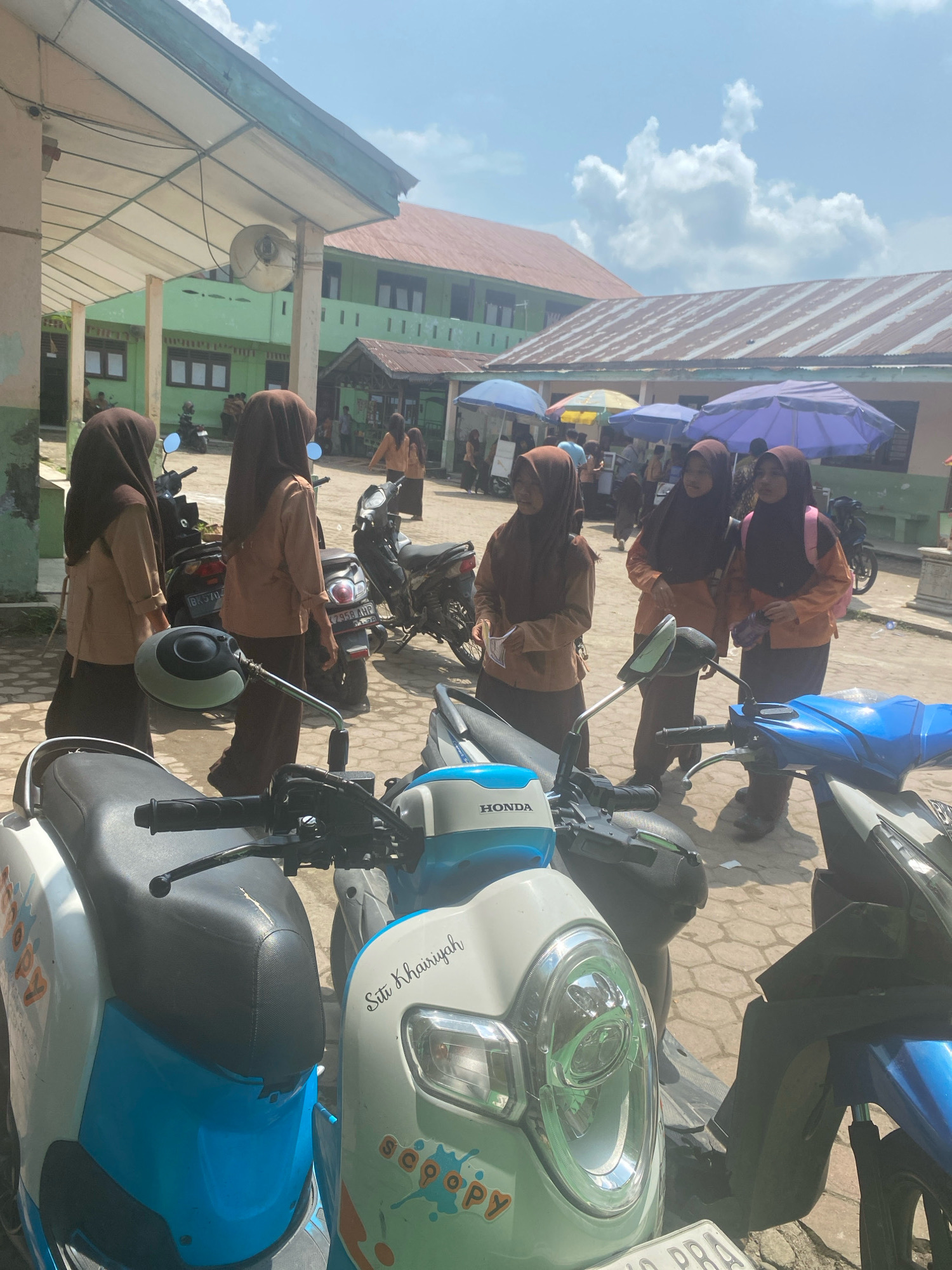 In the city of Karang Gading, Indonesia, a group of young women, likely students, are gathered outdoors in what appears to be a schoolyard. They are wearing uniforms consisting of brown hijabs and light brown tops with dark brown skirts. Several motorcycles are parked around them. In the background, buildings with green walls and reddish-brown roofs are visible, suggesting an educational institution. The sky is blue with scattered white clouds. The overall atmosphere seems casual and relaxed, possibly during a break or after school.
