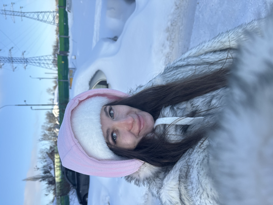 The image presents a forensic view of a female individual in Molodi, Russia, situated in a deeply snow-covered outdoor environment. The subject occupies the foreground, centered, directly facing the camera with a slight smile. She wears a light pink hooded garment with a white furry lining, layered over a grey and white faux fur coat; strands of dark brown hair are visible beneath her hood. Her facial features include light-colored eyes and a rosy complexion. The background reveals a landscape uniformly covered in thick snow under a clear blue sky.

To the subject's left, a dark-colored sedan is partially visible, covered by snow. To her right, a white vehicle is almost entirely buried under snow, with its roofline and windows barely discernible. Further right, another vehicle shape is completely obscured by snow. A distinct green fence or barrier extends horizontally across the mid-ground. Behind this barrier, two large, metallic lattice-style electrical transmission towers stand prominently against the sky. Multiple streetlights are visible throughout the background. Distant structures and sparse, snow-dusted bare trees are also present in the far background. The heavy snow cover indicates severe winter conditions.