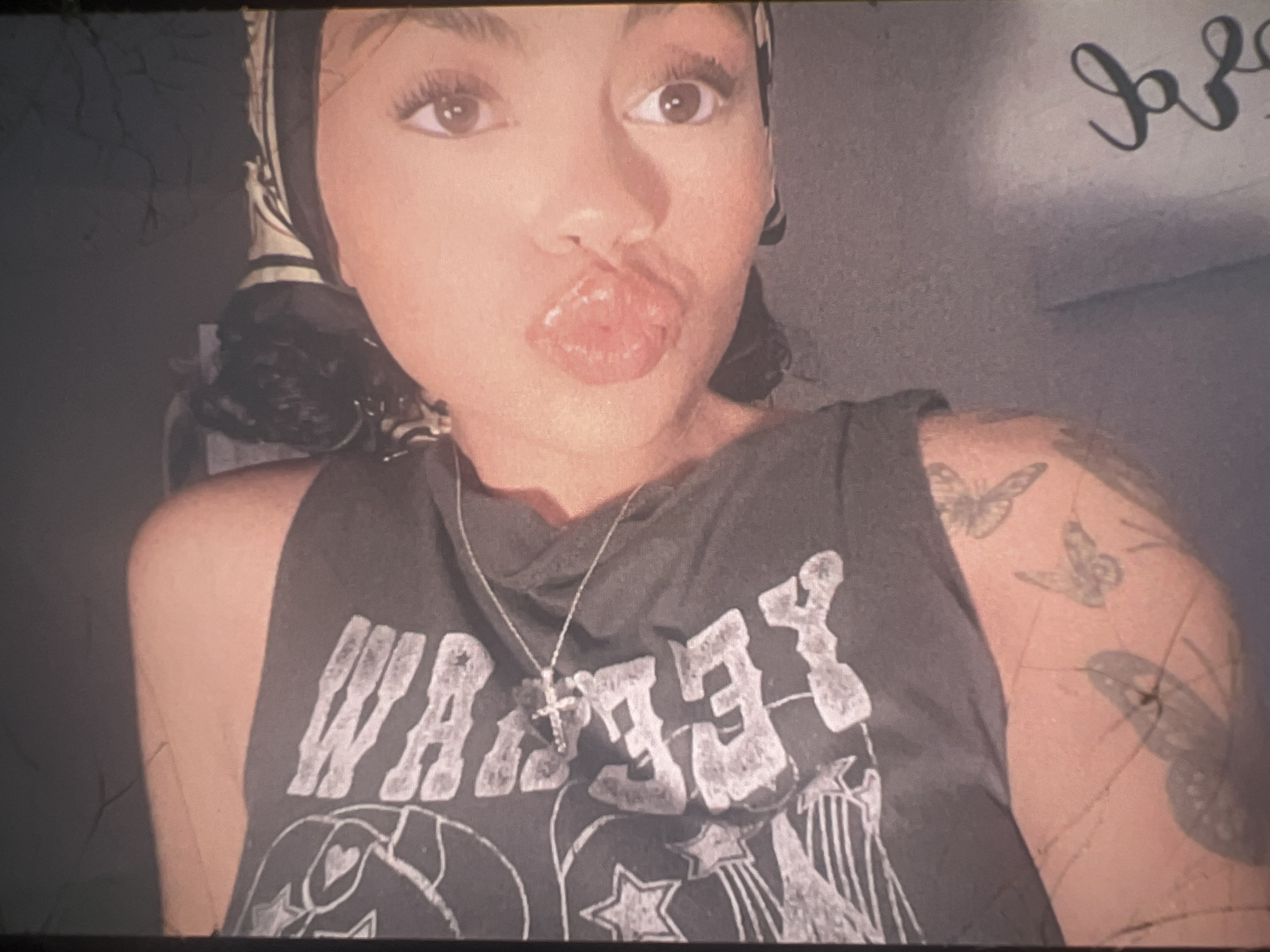 The image is a close-up, rotated selfie of a person lying down, with their face close to the camera. The person has dark hair pulled back and is wearing a dark t-shirt with a white graphic design, a necklace with a cross pendant, and a bandana or scarf around their head with a striped pattern. Their lips are pursed, and their eyes are looking towards the camera. There are tattoos of butterflies on the person's arm, visible on the left side of the image. The background appears to be a gray surface, possibly a bed or sofa. There is no identifiable information about the location in the city of Boone, United States of America.