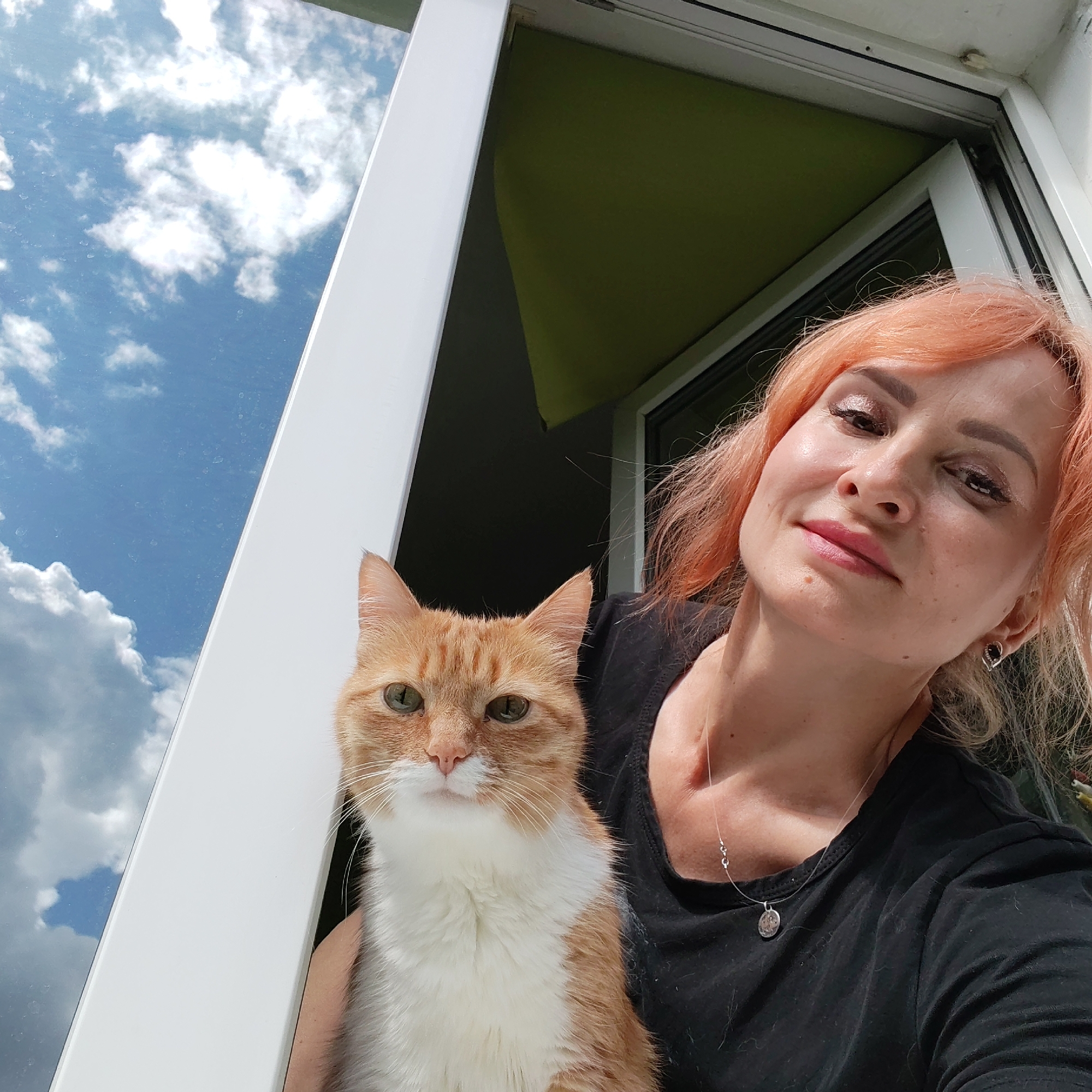 The image depicts a woman with orange hair holding an orange and white cat in front of a window. The woman is wearing a black shirt and a necklace. The cat has green eyes. Outside the window, there is a blue sky with white clouds and a green awning. The scene appears to be on a balcony or in a room with a view of the sky. The context of Chaikovsky, Russia, is not ascertainable from the image content.