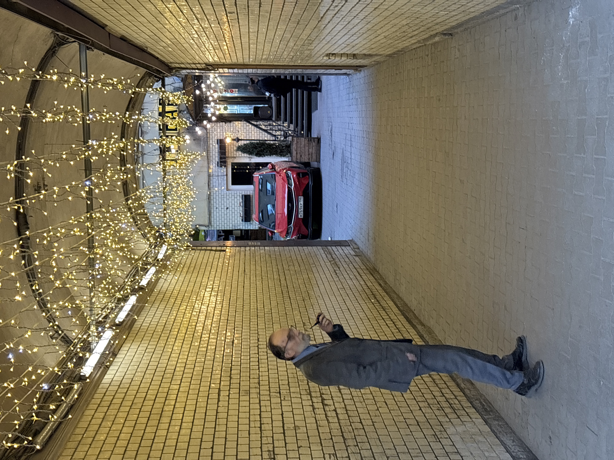 The image captures a man standing within a tunnel-like structure in Saint Petersburg, Russia. He is holding and smoking a pipe, wearing a gray suit and dark shoes, and is positioned against a wall tiled with yellow brick. Overhead, the tunnel's ceiling is lined with strings of small, warm-toned lights.
At the tunnel's end, an exterior scene unfolds, revealing a red car parked in front of a building that appears to be a restaurant or bar, suggested by the "Cafe" sign. Additional figures are present near the entrance of the establishment. The exterior is illuminated by decorative lighting as well. The tunnel itself is constructed of brick.