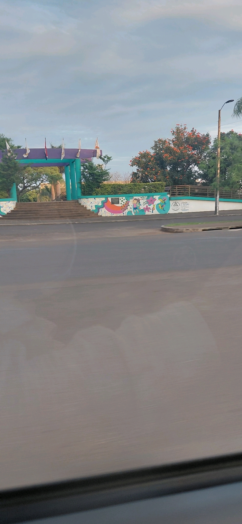 The image depicts an entrance archway with multiple flags on top, situated in Encarnación, Paraguay. The archway is painted in vibrant colors, predominantly purple and teal, and is flanked by decorative murals on a white wall. The murals feature colorful designs including flowers, birds, and a child. Steps lead up to the archway, which is surrounded by lush green trees and foliage. A street is visible in the foreground, suggesting the entrance is accessible from a road. A street lamp is also present to the right of the scene. The sky is overcast with a light blue hue.