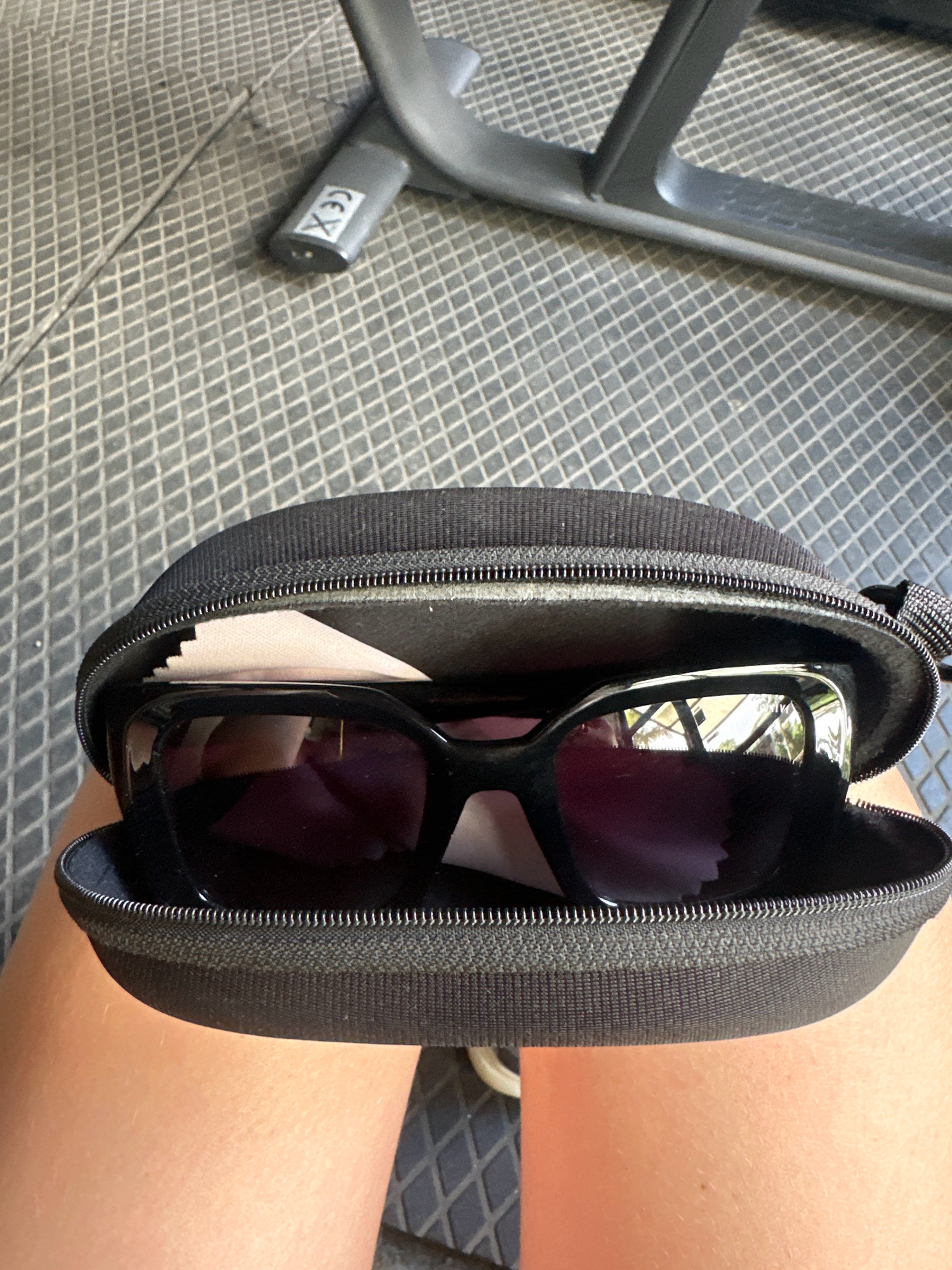 The image shows a pair of black sunglasses with purple lenses inside an open black zippered sunglasses case. A light-colored cloth is placed on top of the sunglasses, likely for protection. The case is resting on a textured gray surface, possibly a mat or flooring. In the background, part of a gray metal exercise machine is visible, and a circular stamp with "CE" is present on one of its components. The image is taken from a slightly elevated angle, focusing on the sunglasses and their case. The location in Lalitpur, Nepal, is not visually represented in the image itself.