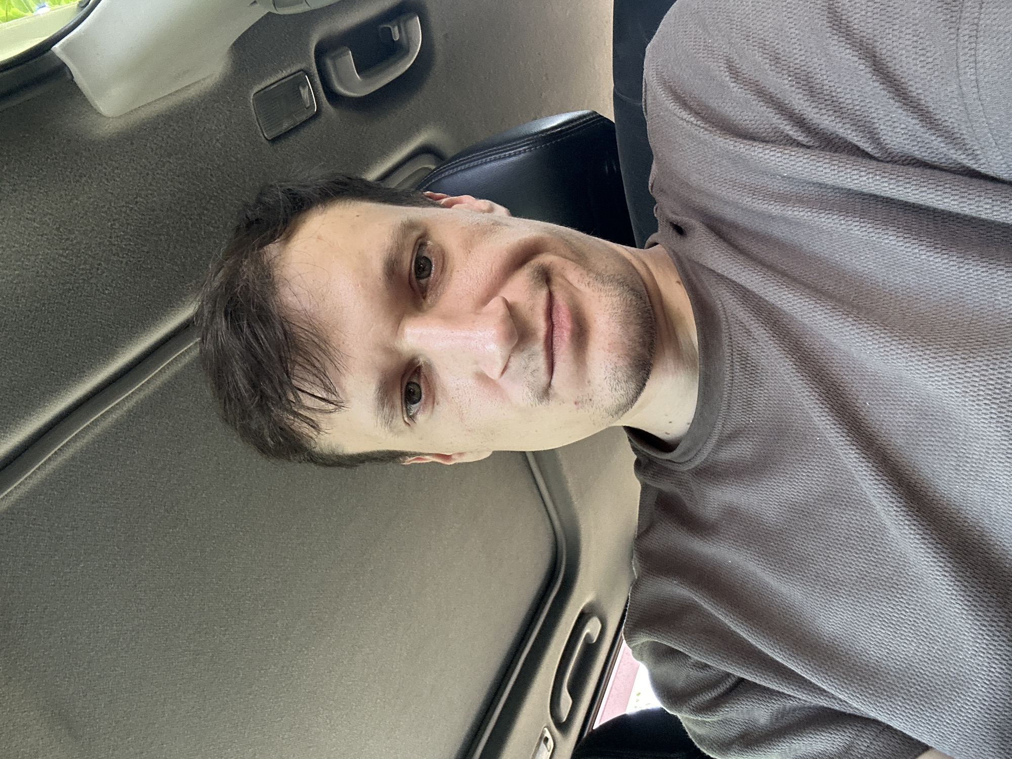 A man is seated inside a vehicle, facing the camera. He has short, dark brown hair and is wearing a brown, textured t-shirt. The interior of the car is visible, including a sunroof and a black seat. The lighting suggests it is daytime. The man's facial expression is neutral.