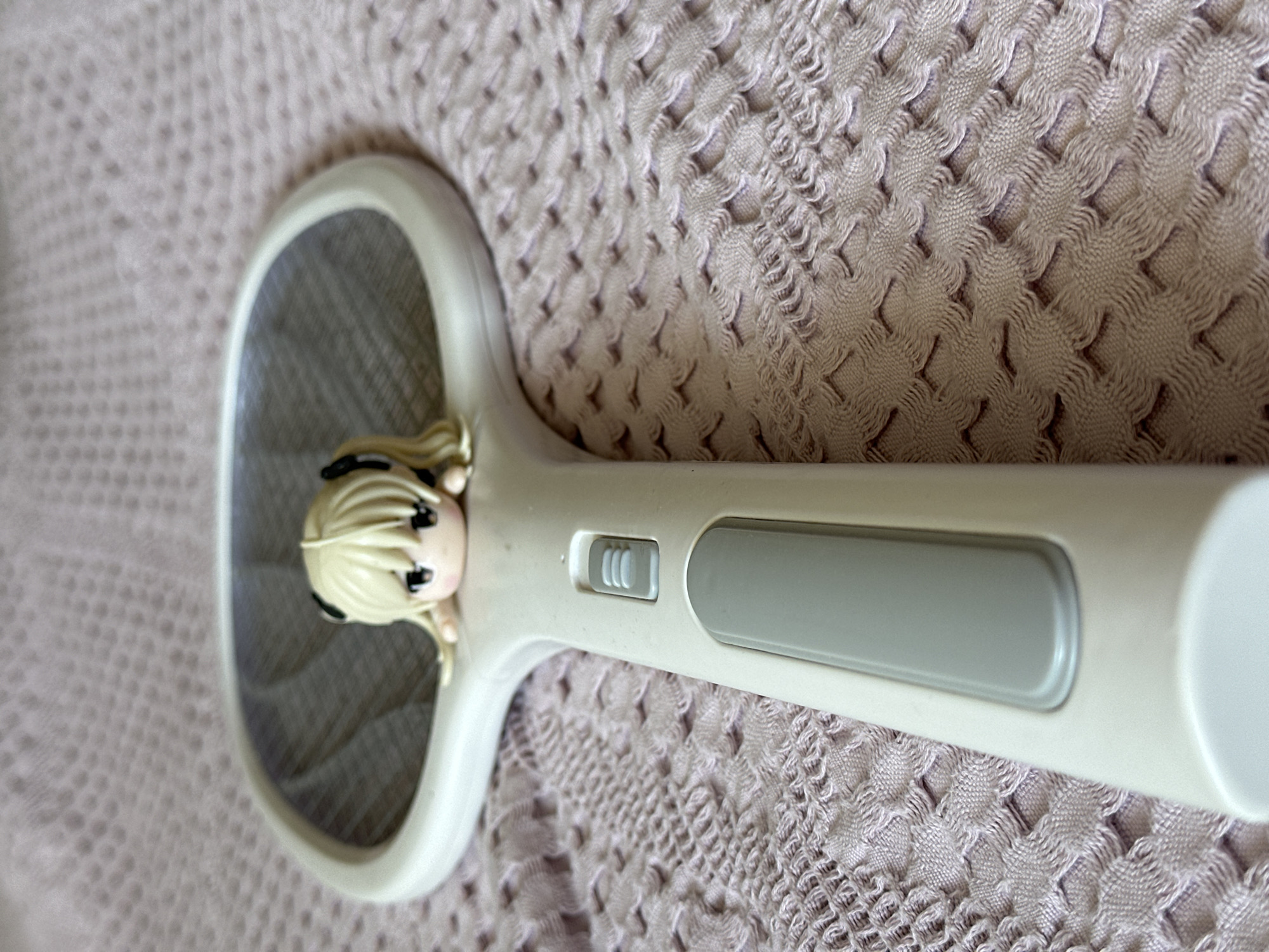 A close-up, low-angle shot focuses on a white electric fly swatter placed on a pink, textured fabric. Resting on the edge of the swatter's mesh head is a small, blonde anime figurine with black-tied pigtails and large, dark eyes. The handle of the fly swatter, which has a grey button and a larger grey grip, extends downwards and out of frame. The fabric appears to be a blanket or throw with a repetitive, intricate weave pattern. The lighting suggests an indoor setting. This image has no identifiable elements placing it in Nevinnomyssk, Russia.