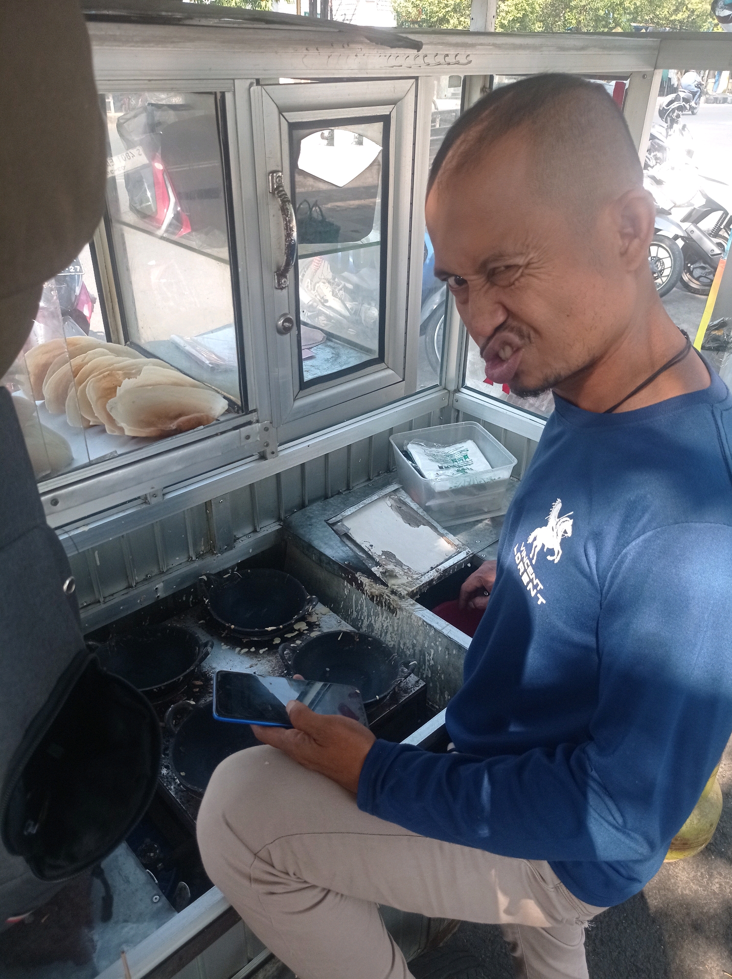 A man with a shaved head and a grimace is leaning over a food cart in Tuban, Indonesia. He is wearing a blue long-sleeved shirt with a white horse and rider logo and the words "VINCENT LORENT" printed on it. He is holding a smartphone in his right hand. The food cart has a glass display case with a stack of round, flat pastries, and a cooking area with several black circular pans. In the background, motorcycles are visible on a street.