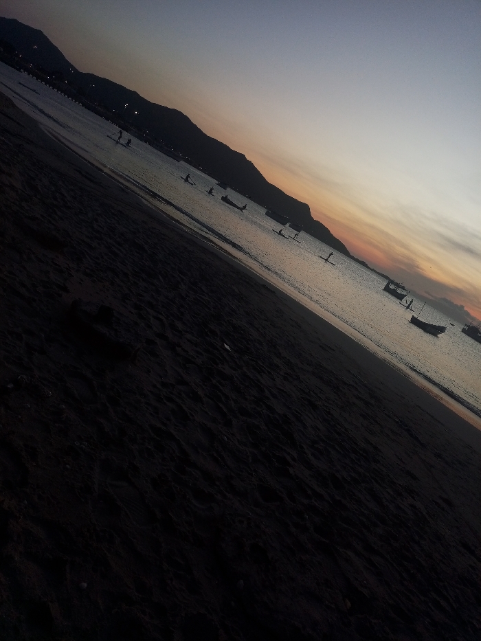A dark, angled view shows a beach at dusk or dawn. Mountains line the horizon under a sky transitioning from purple to orange. Silhouettes of people stand on paddleboards in the water, alongside several boats anchored near the shore. The foreground consists of dark sand, creating a stark contrast to the lighter sky and water.