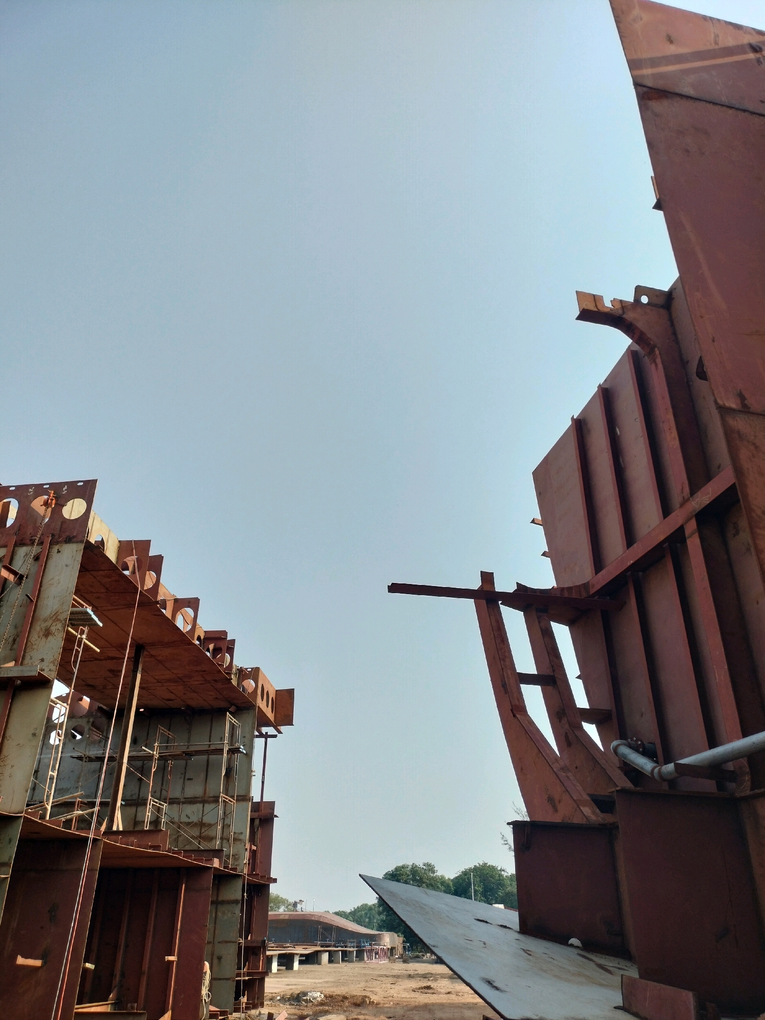 This is a low-angle, full shot of a shipyard in Batam, Indonesia, showcasing the construction of large metal structures, likely ship hulls or sections of vessels. The sky is clear and light blue, taking up most of the upper portion of the frame.
On the left, a towering, complex metal structure is in progress. It features numerous beams, plates, and vertical supports, some with circular cutouts. Scaffolding is visible attached to the interior of this structure. A chain hoist is suspended from one of the beams. The metal is a rusty, reddish-brown color.
On the right, another massive, rust-colored metal structure dominates the foreground. It has a curved, angular shape with thick beams and large plates forming its exterior. A silver pipe runs horizontally along the top of this structure.
In the background, beyond the immediate construction site, there are more structures, some appearing to be under construction or completed sections of vessels. Trees and vegetation are visible in the distance, suggesting a coastal or industrial area. The ground appears to be dirt or gravel, consistent with a construction environment. No people are clearly visible in the image, though some faint figures might be present in the distance. The overall impression is one of large-scale industrial construction.