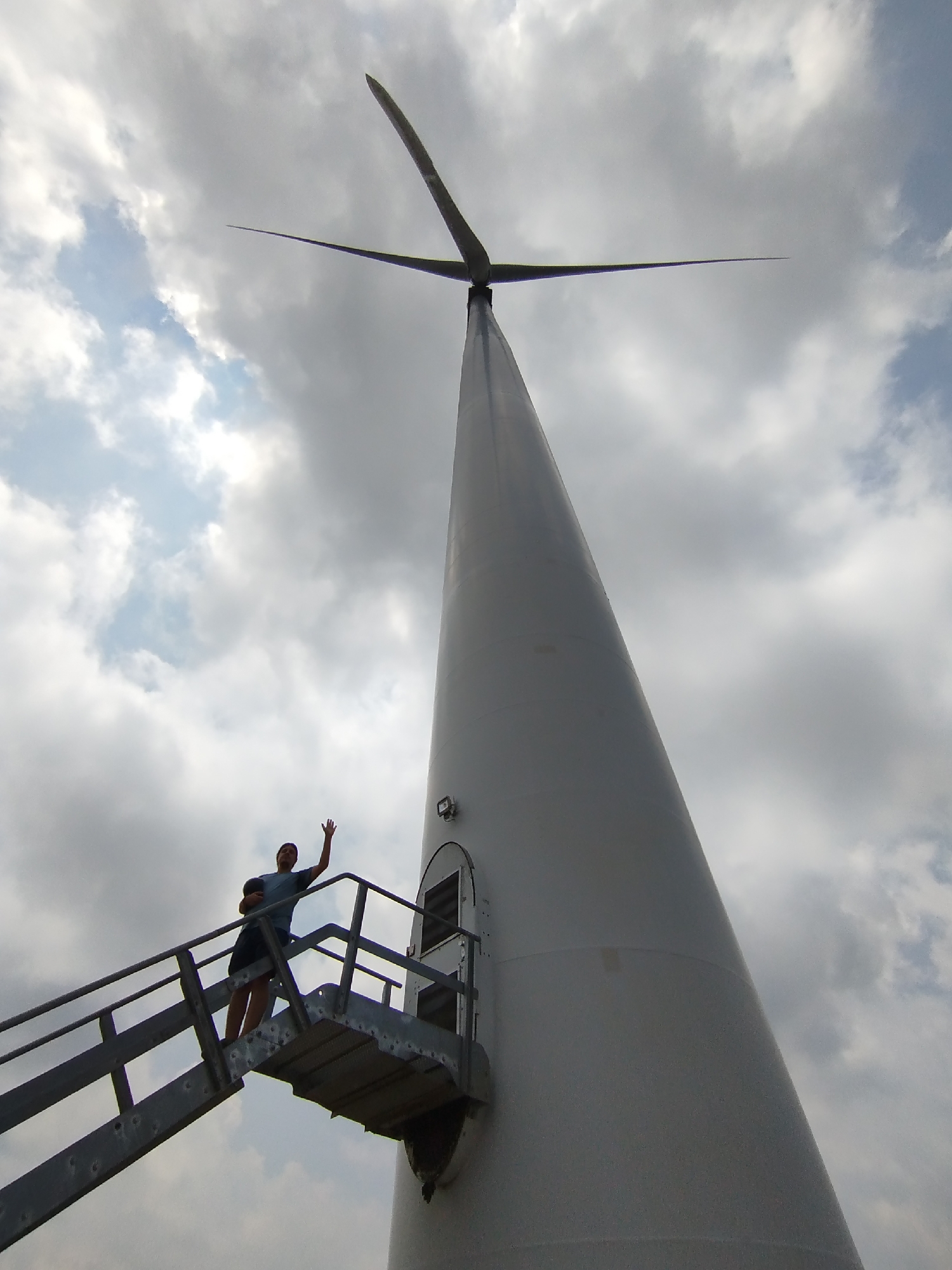 A low-angle shot shows a wind turbine with a man standing on a platform near its base, waving. The man is wearing a t-shirt and shorts. He is standing on a metal platform with a railing, part of a staircase leading to a door in the turbine. The turbine tower is white and tapers upward, ending in a three-blade rotor. The sky is filled with cloudy formations. The location is Phát Chi, Vietnam.