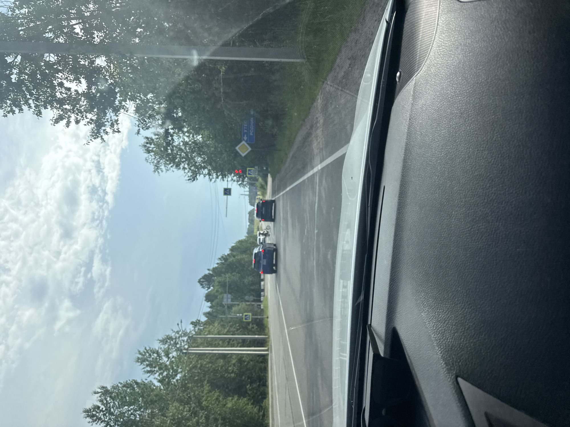 The image is taken from inside a car, looking out at a road in Russia. The road is asphalt and has white lane markings. There are trees and foliage on either side of the road, and the sky is visible, with some clouds. The time of day appears to be daytime, possibly late morning or early afternoon, given the bright sunlight.
Several cars are on the road, moving in the same direction as the camera car. The closest car to the camera is a dark-colored SUV, followed by two other dark-colored cars. A motorcycle is also visible on the road ahead.
There are several road signs visible. One sign, with blue lettering, points to "УТКБ РОДНИК" (UTKB Rodnik). Another sign is a yellow diamond shape, likely indicating a warning. There are also traffic lights, which appear to be red. A pedestrian crossing sign is also visible.
The overall scene suggests a typical drive through a rural or suburban area in Russia. The weather appears to be clear and sunny. The interior of the car, specifically the dashboard on the right side of the image, is also partially visible, showing textured black plastic.