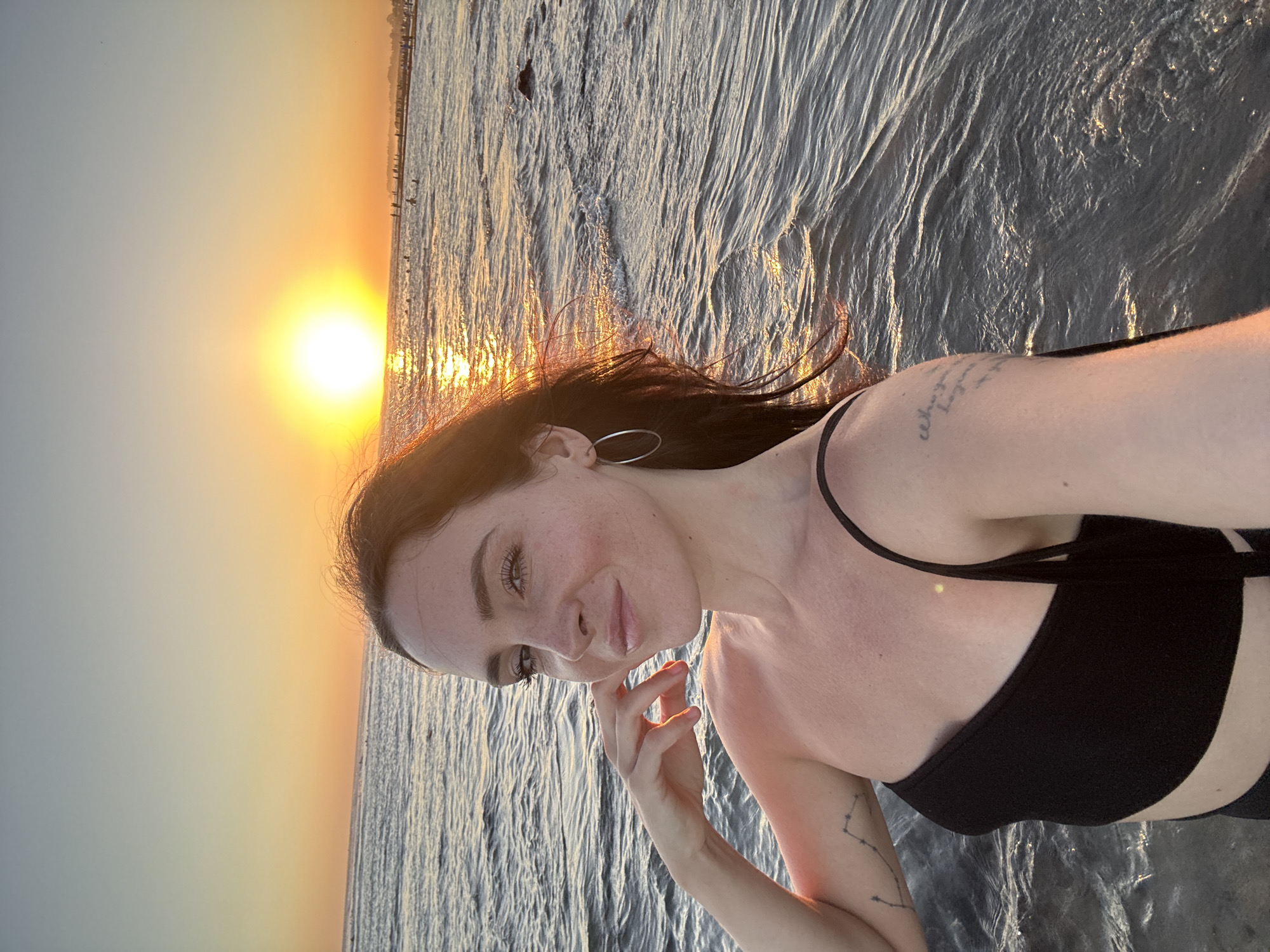 A woman is taking a selfie in the ocean at sunset in North Kuta, Indonesia. She is wearing a black bikini top and has her hand on her chin. Her hair is dark and wavy, and she is smiling at the camera. The sun is setting behind her, casting a warm glow over the water. There are other people in the background, enjoying the beach. The waves are gentle, and the water is clear. The overall atmosphere is one of peace and relaxation.