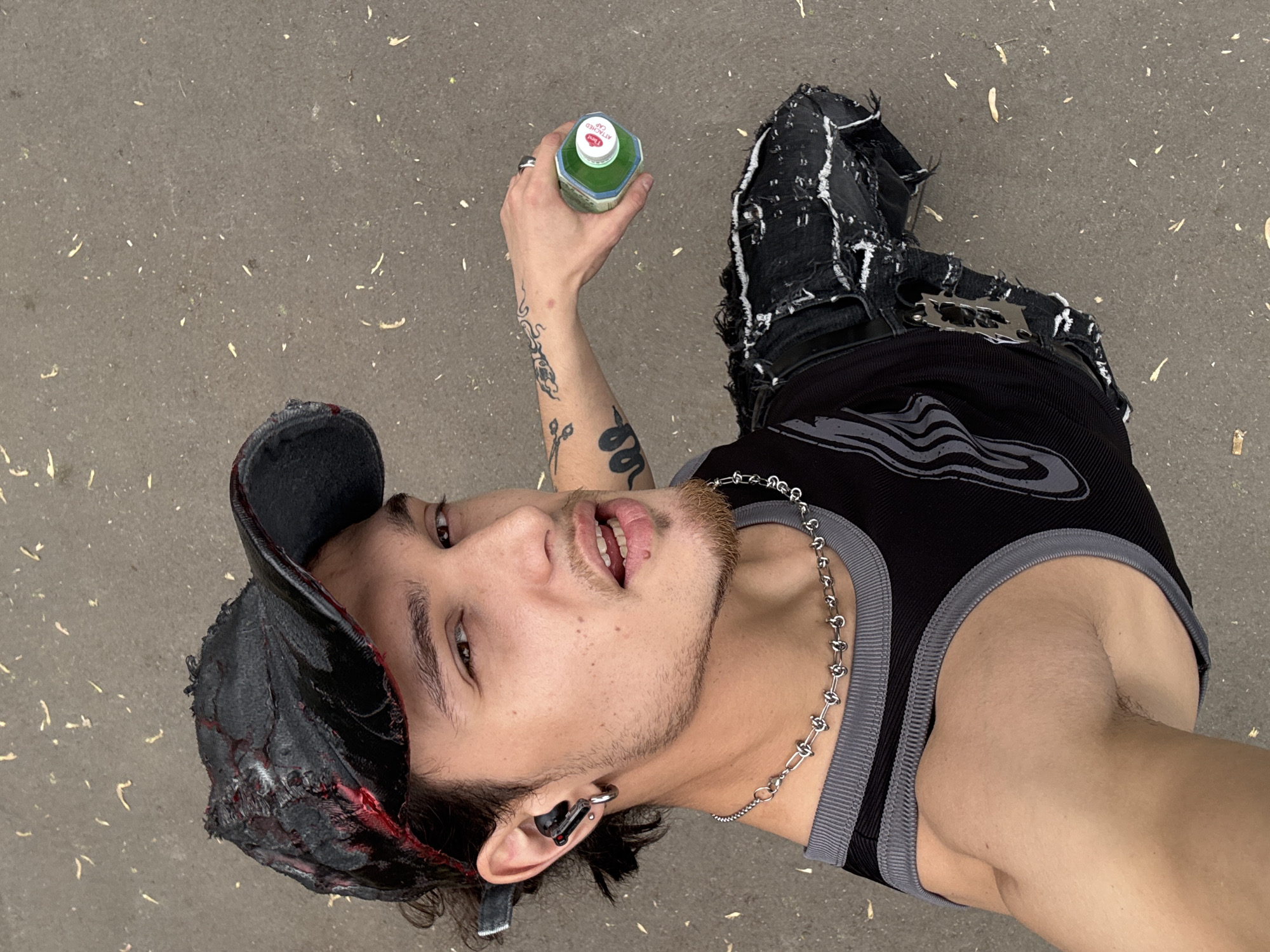 A medium overhead shot depicts a young man wearing a black distressed cap, black tank top with gray accents, distressed black jeans, and a silver chain necklace. He is holding a green bottle in his right hand. Tattoos are visible on his right forearm. The scene is set on a concrete ground. The location cannot be identified as Sofia, Bulgaria, from the visual information.