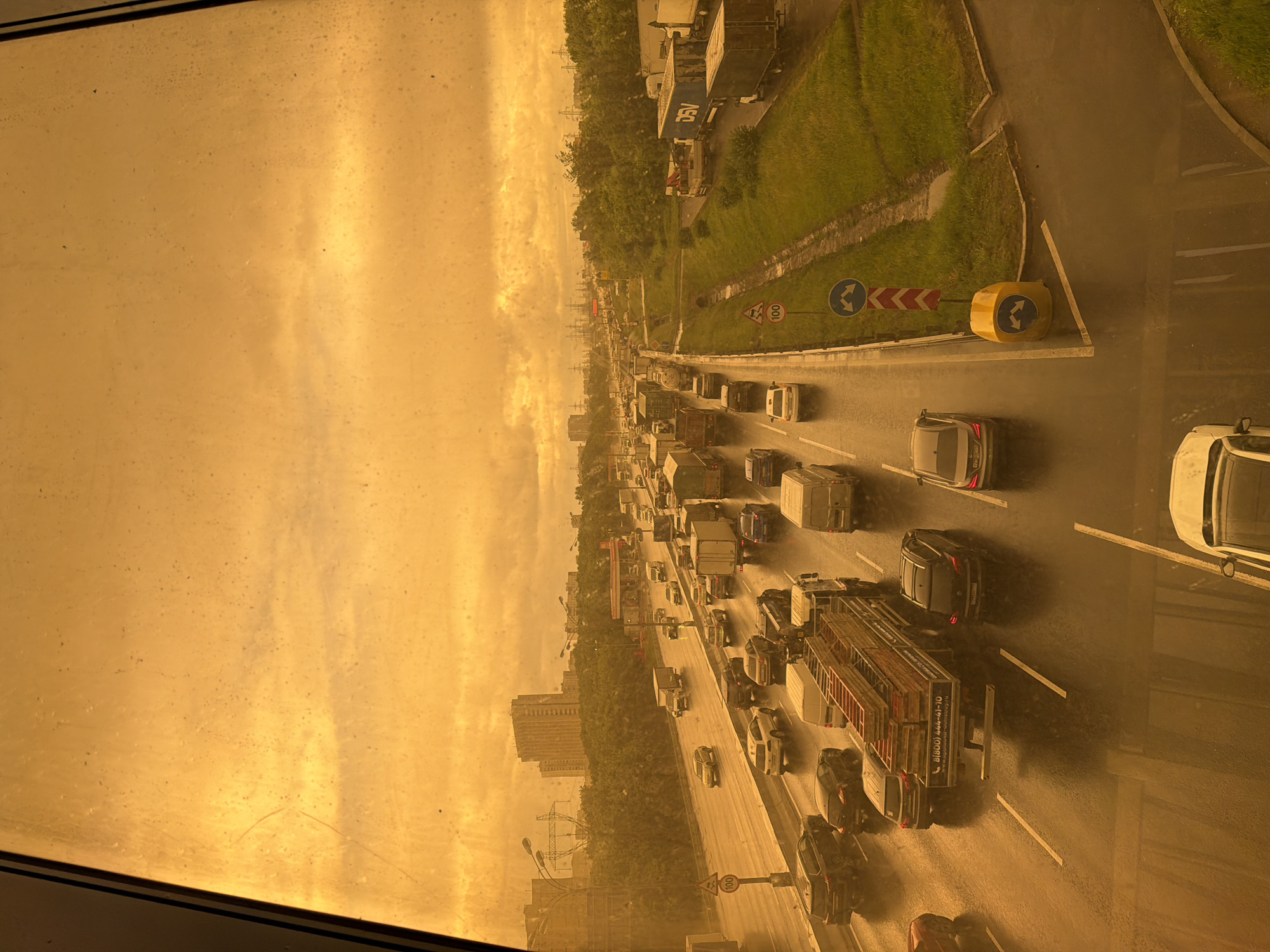 The image depicts a traffic jam on a multi-lane highway in an urban area. Numerous vehicles, including cars and trucks, are visible on the roadway. The sky is a hazy yellow, potentially indicating atmospheric conditions such as dust or pollution. The view is from an elevated position, possibly a window in a building overlooking the highway. There are buildings and greenery along the side of the road. Signage is visible along the side of the highway, including road signs indicating a curve and speed limits.