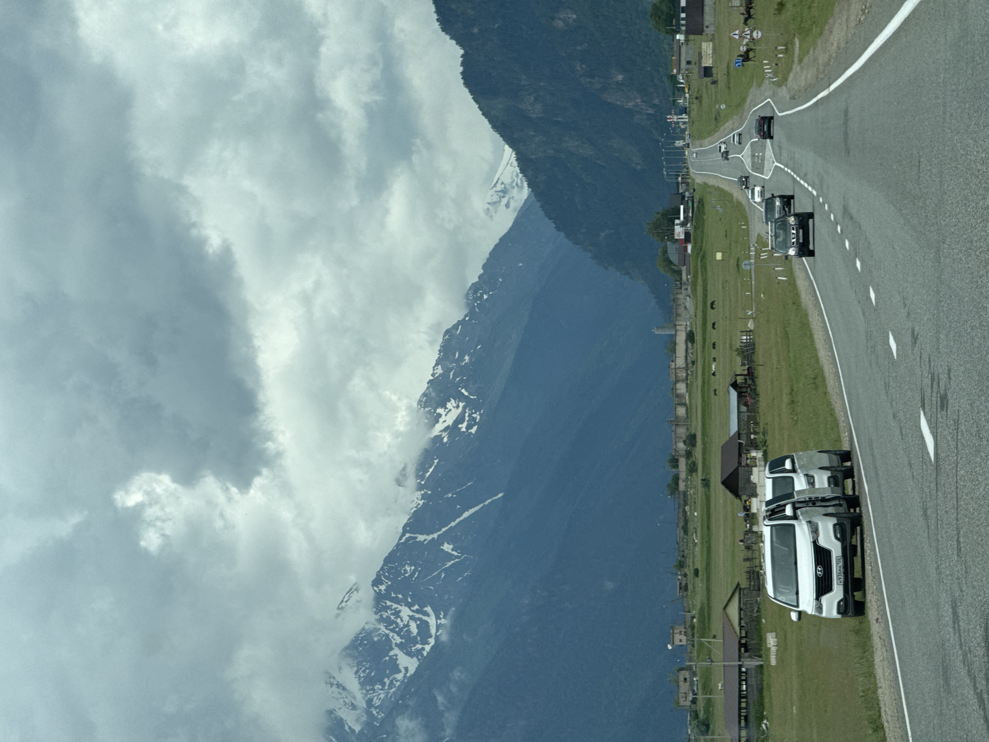 The image captures a scenic view of a road leading into a mountainous region near the city of Verkhnyaya Teberda, Russia. The foreground features a paved road with white lane markings, and on the shoulder, two white passenger vans are parked with their doors open. Several other vehicles, including cars and SUVs, are traveling along the road in both directions. The middle ground displays a vast green field dotted with scattered buildings, some appearing to be farm structures and houses. A herd of animals, possibly horses, is visible grazing in the field. In the background, imposing, dark blue mountains rise dramatically, their upper reaches capped with snow and shrouded in cloudy mist. The sky is overcast with heavy, grey clouds, suggesting potentially cool or inclement weather. The overall impression is one of a rural, mountainous landscape with a road serving as a connection to a remote settlement.