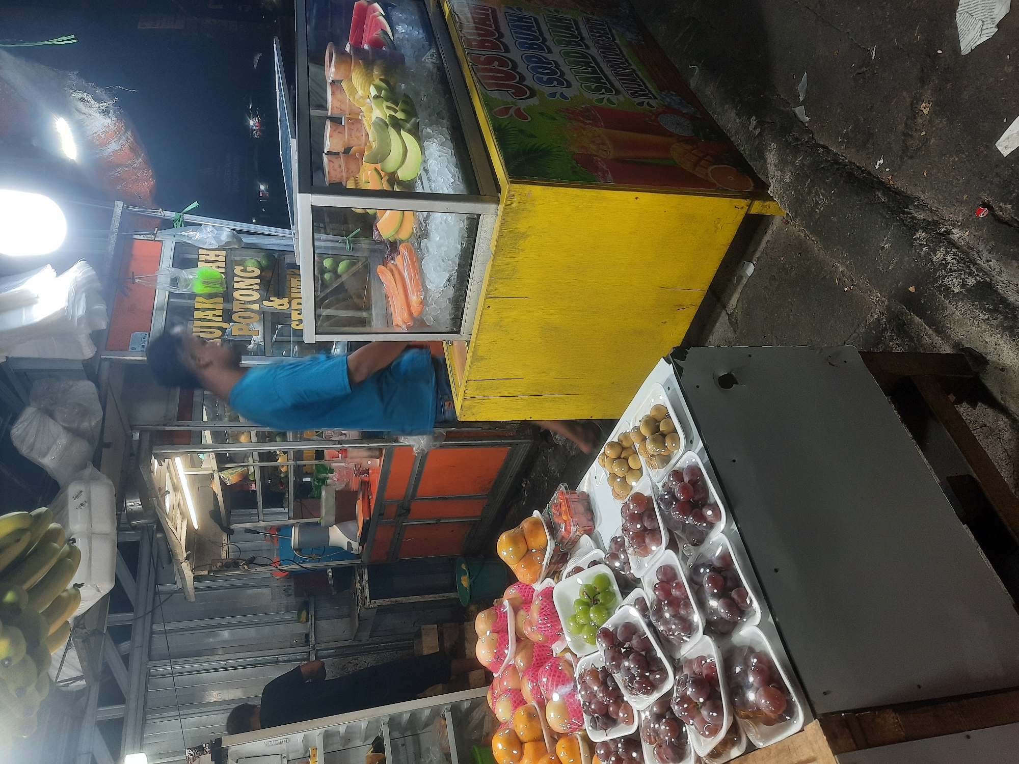 At night, a fruit stand in North Jakarta, Indonesia, is illuminated by artificial lights. The stand is filled with various fruits, including grapes, longans, peaches, oranges, and watermelon, displayed in plastic packaging on trays. A refrigerated glass display case showcases sliced fruits like cantaloupe, honeydew melon, and pineapple, on a bed of ice. Behind the display, a yellow wooden structure features colorful signage for "JUS BUAH SOP BUAH SALAD BUAH RUJAK POTONG SERUT". Two individuals are partially visible; one, wearing a blue t-shirt, is in the process of serving customers, while another, wearing a black t-shirt, stands near a refrigerator. The ground is a wet, dark asphalt surface, with some debris present. The background suggests a street environment with metal structures and some overhead awnings.