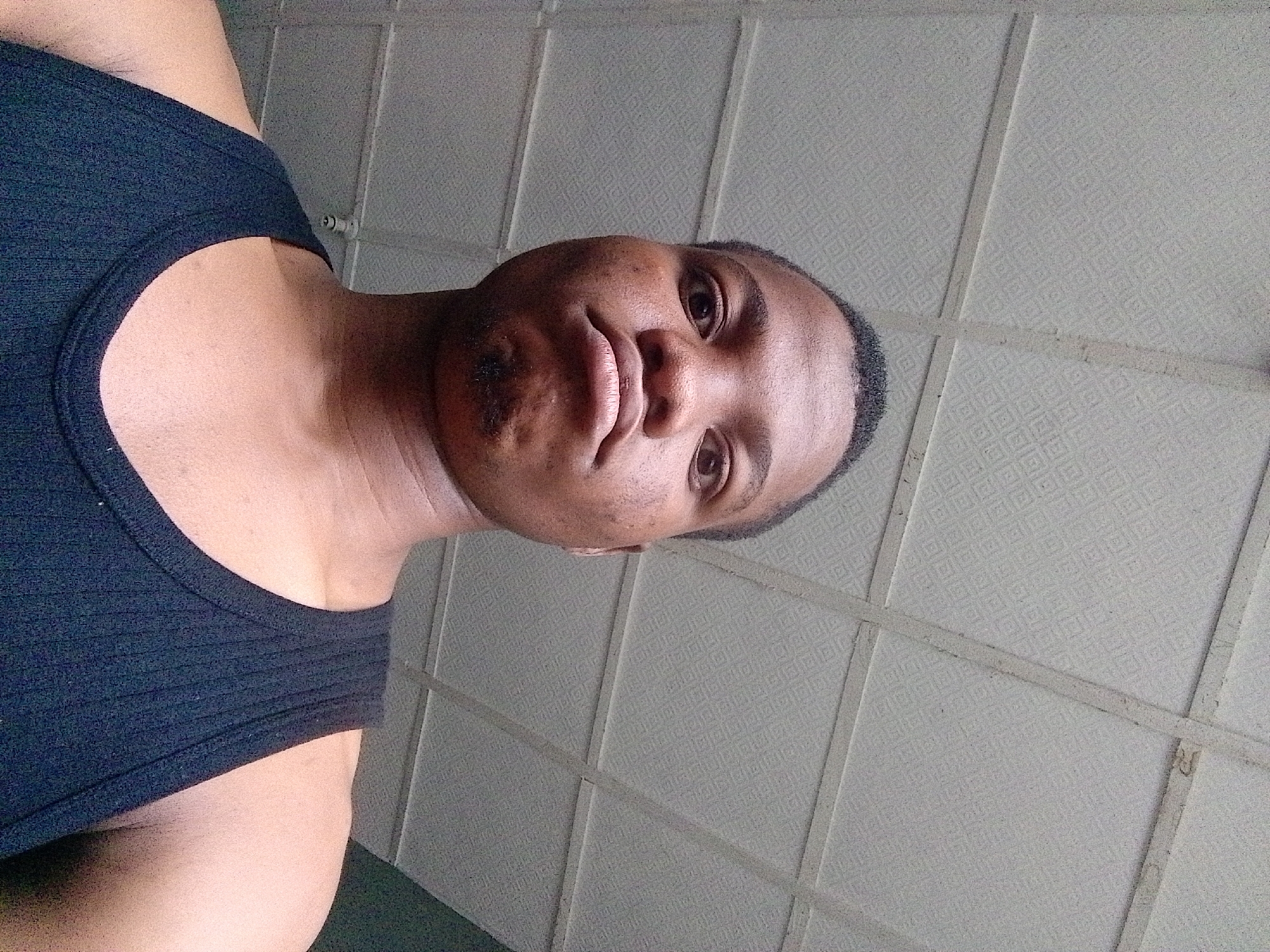 A close-up, low-angle, full-body, vertical, outdoor, candid shot shows a person from the chest up, facing the camera. The person is looking directly at the camera with their head slightly tilted. They have dark skin, short dark hair, and are wearing a black tank top. The background is a light gray, textured ceiling with visible seams. The lighting appears to be natural, coming from overhead. The scene is indoors, possibly in a residential or public space. There is no indication of the specific city or location.