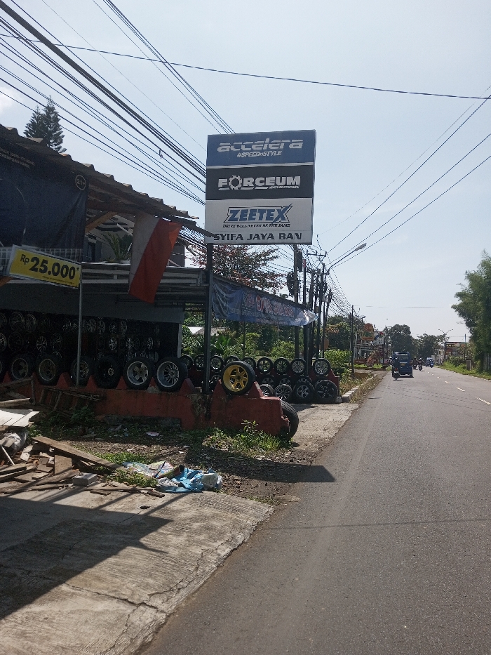 The image presents an outdoor commercial scene under clear, bright daylight in Beteng_Pangkhar_Somthrang, Bhutan. In the left foreground, a cracked concrete sidewalk is strewn with wooden debris and a crumpled blue plastic or fabric item. This leads to a single-story building, an apparent tire and wheel shop, identified by a prominent vertical sign displaying "ACCELERA," "FORCEUM," "ZEETEX," and "SYIFA JAYA BAN." The shop's open facade showcases numerous vehicle tires and alloy wheels, arranged on display stands and a low, red concrete structure. A red and white bicolour flag hangs vertically from the building's overhang, adjacent to a yellow sign indicating "Rp 25.000*." Numerous overhead utility lines crisscross the expanse of the light blue sky. To the right, an asphalt road extends into the background, where a dark sport utility vehicle and a smaller unidentified vehicle are observed traveling away from the camera. Sparse green foliage and trees line the far side of the road, with additional utility poles and wiring visible. No individuals are clearly discernible in the scene. Strong shadows are cast, indicative of direct sunlight.
