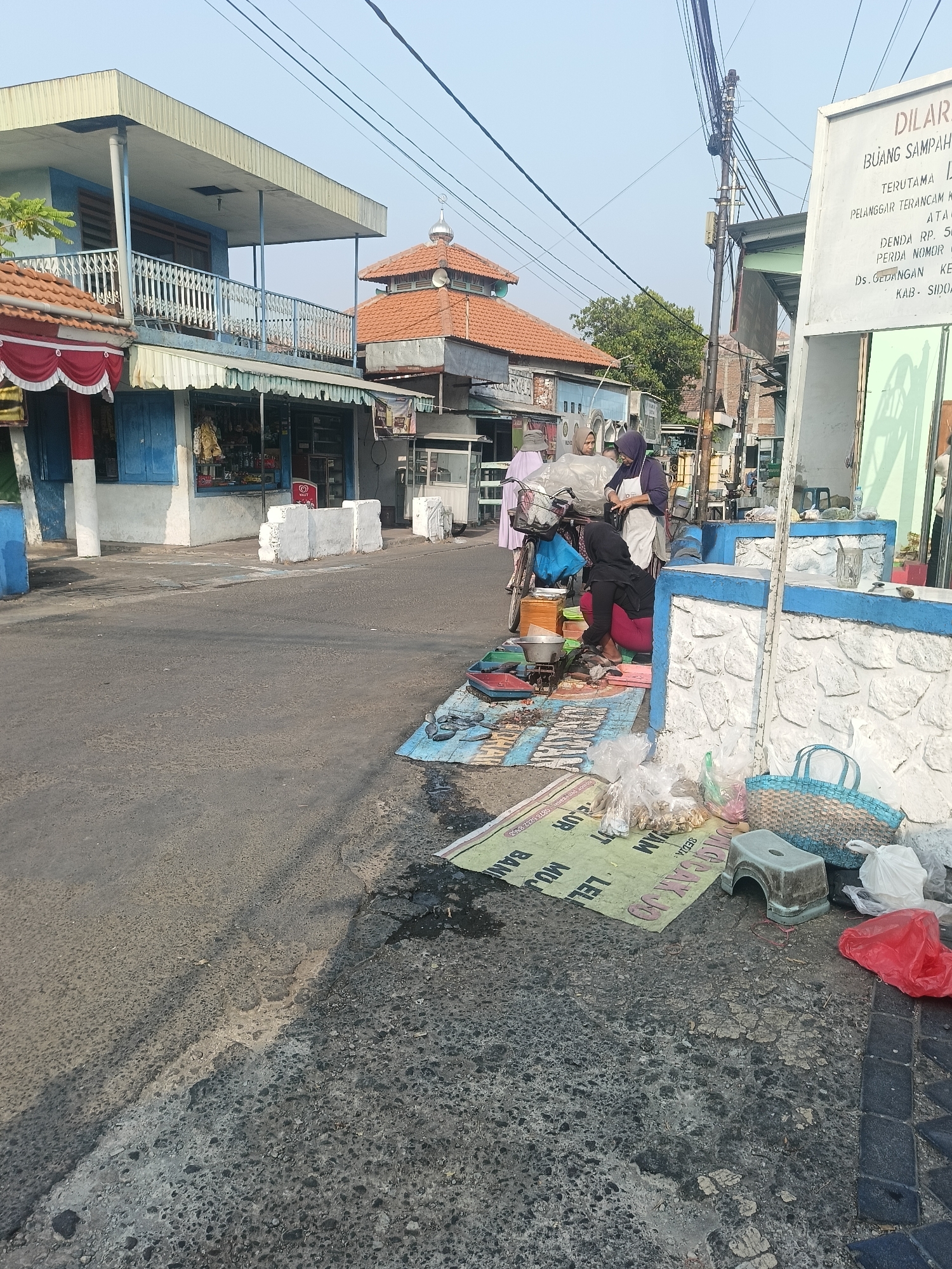The image shows a street scene in Gedangan, Indonesia, with a mosque in the background. In the foreground, a woman wearing a headscarf and apron is seated on the ground next to a bicycle, arranging items on a mat. Another woman, also wearing a headscarf and a light-colored robe, stands nearby, looking down at the items. A third woman, wearing a pink headscarf and a light-colored outer garment, stands beside the bicycle, seemingly engaged in conversation. The mat on the ground is covered with various items, including plastic bags, packaged goods, and some sort of food items. A blue basket and a small stool are also visible on the mat. The street itself is paved with asphalt, and there are buildings on either side of the road, including a multi-story building with a balcony and shops on the ground floor. A white sign with blue lettering is prominently displayed on the right side of the image, which appears to be a public notice or warning. The sky is clear and blue, indicating a sunny day. The overall scene depicts a busy street market or roadside stall.