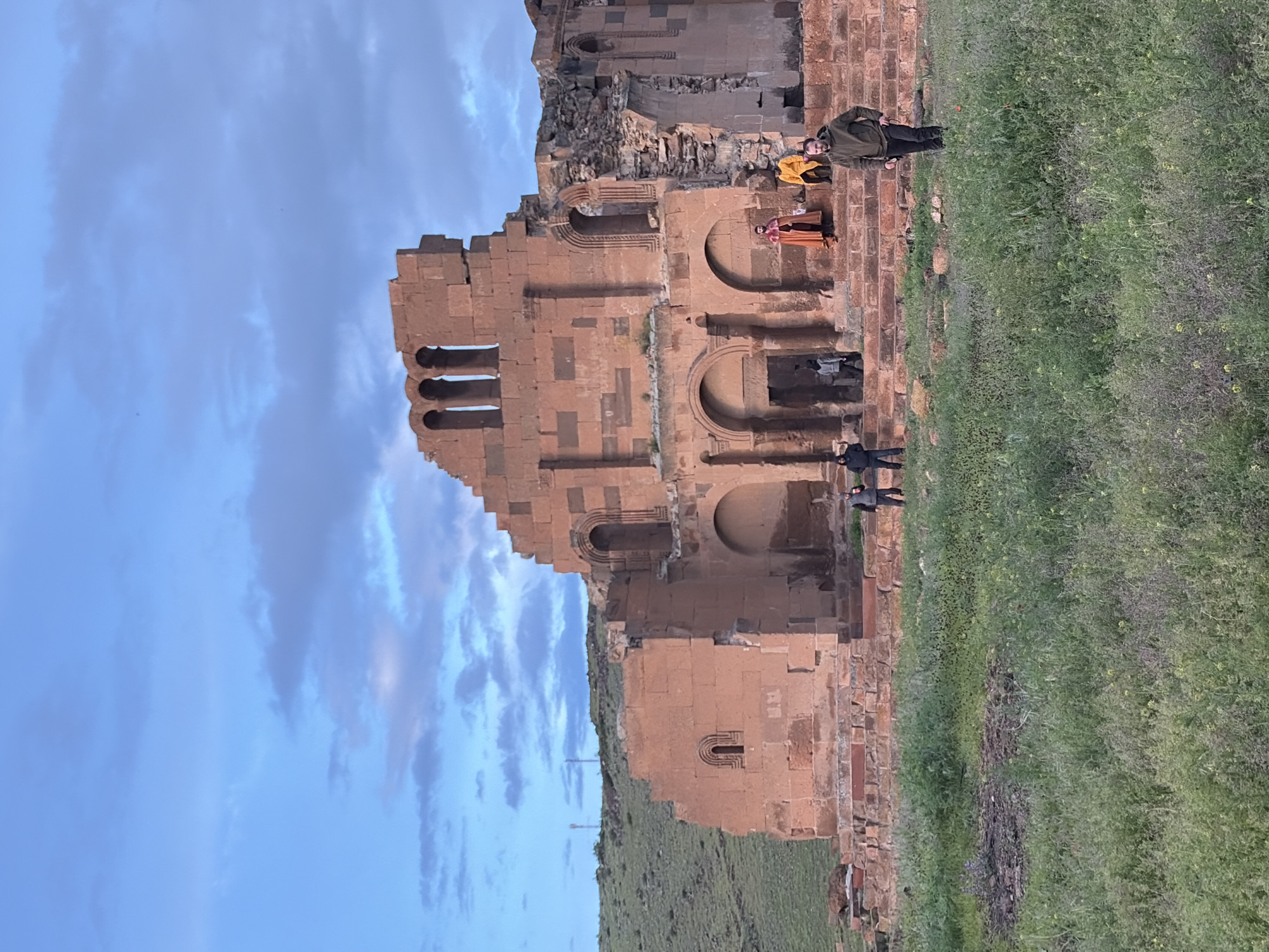 A horizontal, outdoor shot shows the ruins of a red stone structure with people standing in front of it. The structure is located in the city of Anipemza, Armenia. The ruined structure features several arched openings and a row of vertical pillars at its top. To the right of the structure is an expanse of green grass, and to the left is a blue sky with clouds. Several people are visible near the structure: one person is wearing a yellow jacket and another is wearing a red/orange outfit. A person wearing a dark jacket and pants is standing towards the right.