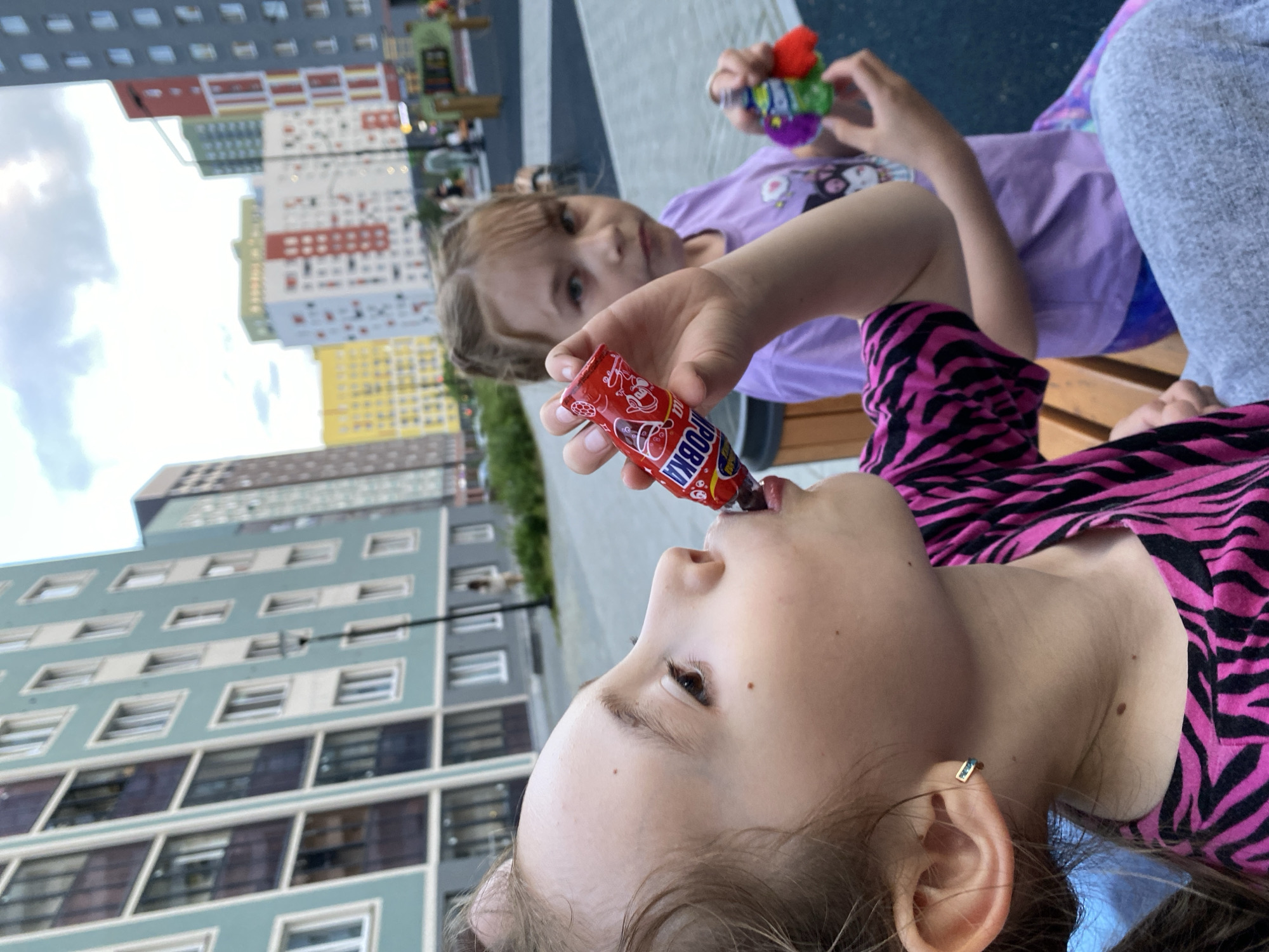 Two young girls are sitting on a bench in a modern residential courtyard in Yekaterinburg, Russia. The girl in the foreground, on the left, is wearing a black and pink zebra-striped shirt and is actively eating a red candy stick. She is captured in a profile view, with her head tilted back as she consumes the candy. The second girl, on the right, is wearing a purple t-shirt with cartoon characters and is holding a small green object with colorful pom-poms in her right hand. Both girls appear to be enjoying their treats and the outdoor environment. The background features tall, multi-colored apartment buildings typical of contemporary Russian urban development, with a cloudy sky overhead. The courtyard itself appears to be paved and includes playground elements.
