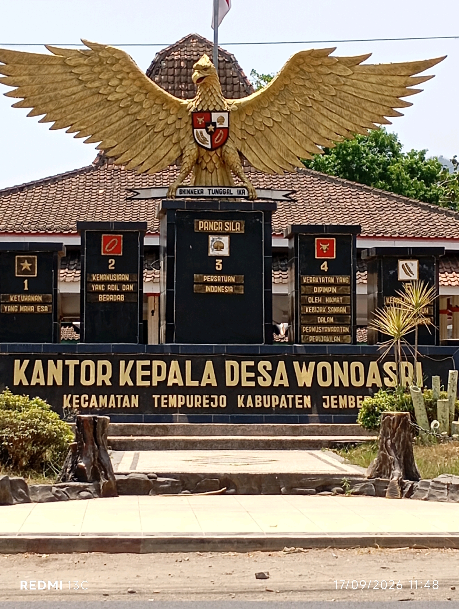 The image shows the exterior of the "KANTOR KEPALA DESA WONOSARI" (Head Office of Wonoasri Village) in the Kecamatan Tempurejo, Kabupaten Jember, Indonesia. Dominating the foreground is a large golden statue of a Garuda bird, the national symbol of Indonesia, with its wings spread wide. Below the Garuda statue is a black monument inscribed with "BHINNEKA TUNGGAL IKA" (Unity in Diversity) and "PANCASILA" (The Five Principles of Indonesian ideology). To the left and right of the central "PANCASILA" inscription are four black pillars, each displaying a number and text relating to the five principles. These pillars are adorned with small emblems, such as a star, a chain, a bull, and a stalk of rice and cotton.
The building behind the monument has a tiled, sloping roof. In front of the building and around the monument, there are stone steps and decorative elements, including what appear to be tree stumps and rocks. Lush green foliage and trees are visible in the background. The sky is bright and clear, suggesting it is daytime. The lower portion of the image shows a street or road with a small stone on it. The bottom right corner displays a timestamp: "17/09/2026 11:48".