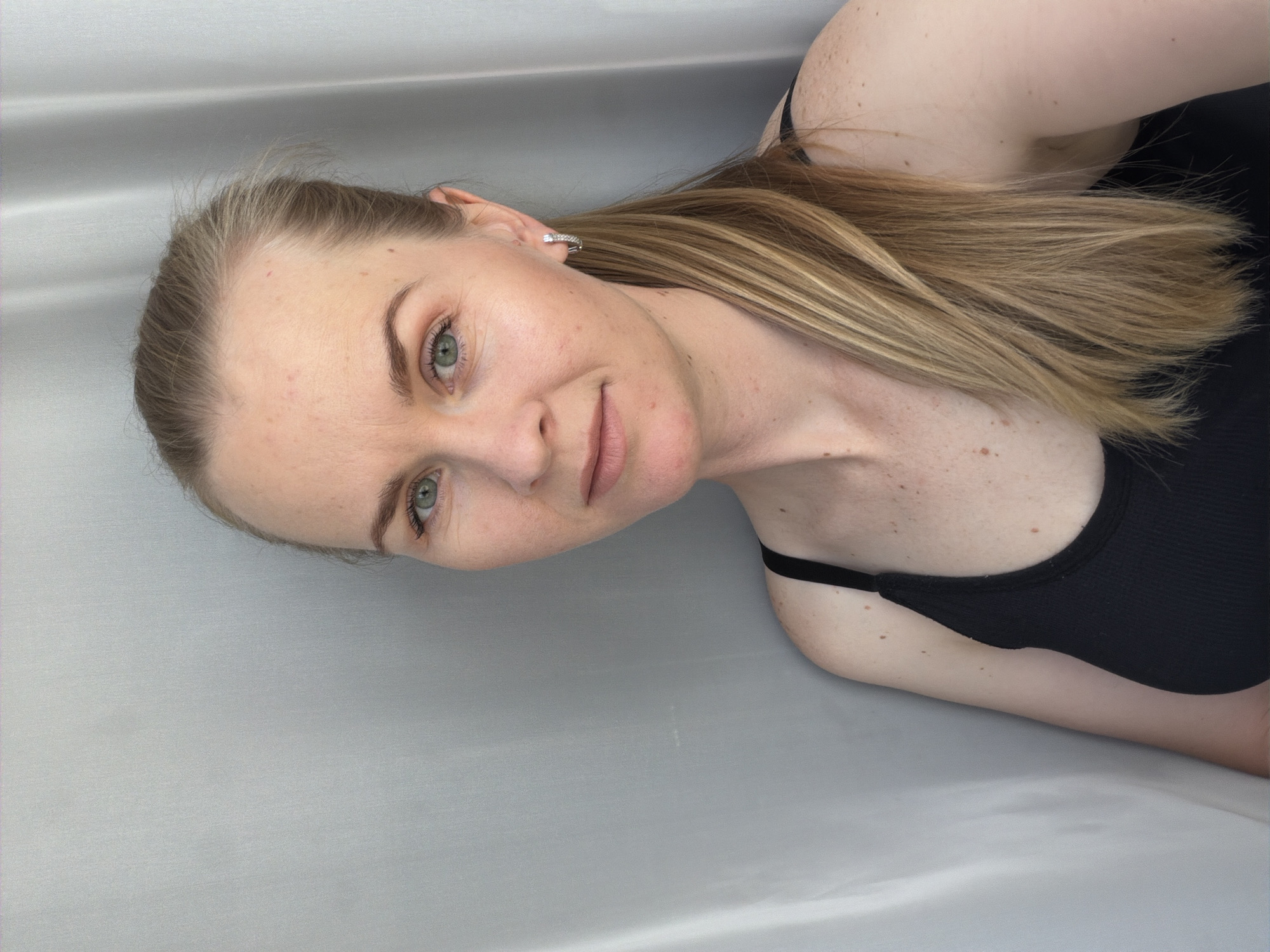 A close-up, head-and-shoulders shot of a fair-skinned woman with blonde hair pulled into a ponytail, wearing a black tank top, and silver hoop earrings. She has light green eyes and freckles scattered across her cheeks and shoulders. The background consists of a neutral, grey-toned fabric that appears to be a curtain or backdrop. The lighting is soft and diffused, creating a natural look. The woman is positioned slightly to the left of the frame, looking directly at the camera with a neutral expression. The environment suggests an indoor setting, likely a studio or a well-lit room. There are no visible contextual clues to indicate the specific location within the city of Кульдур, Russia.